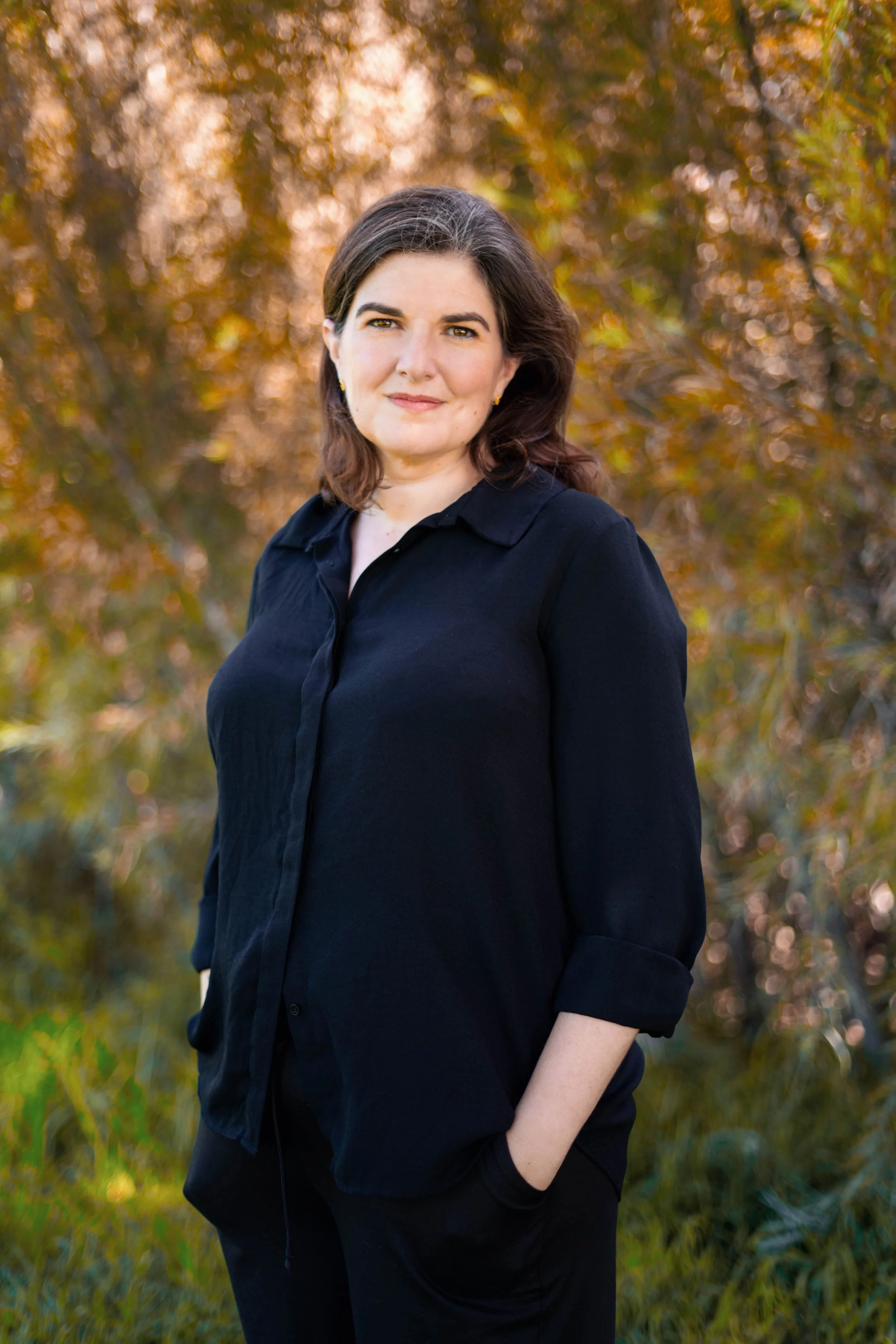 A woman with dark hair wearing a black shirt and black pants stands outdoors with her hands in her pockets, in front of trees with autumn-colored leaves.