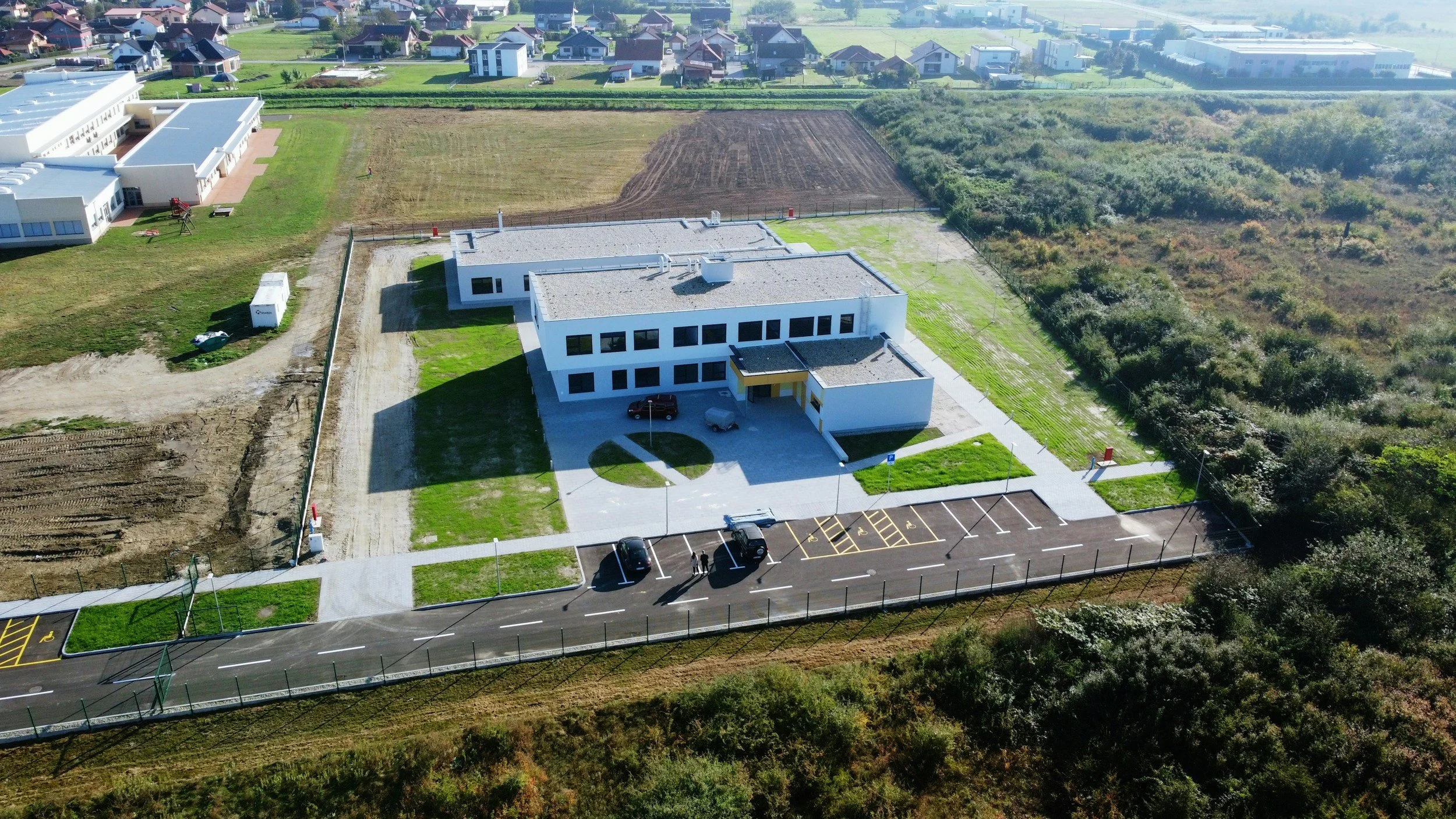 An aerial view of a modern, white, multi-story building with a parking lot and landscaped surroundings. In the background, there are residential houses, green fields, and wooded areas.