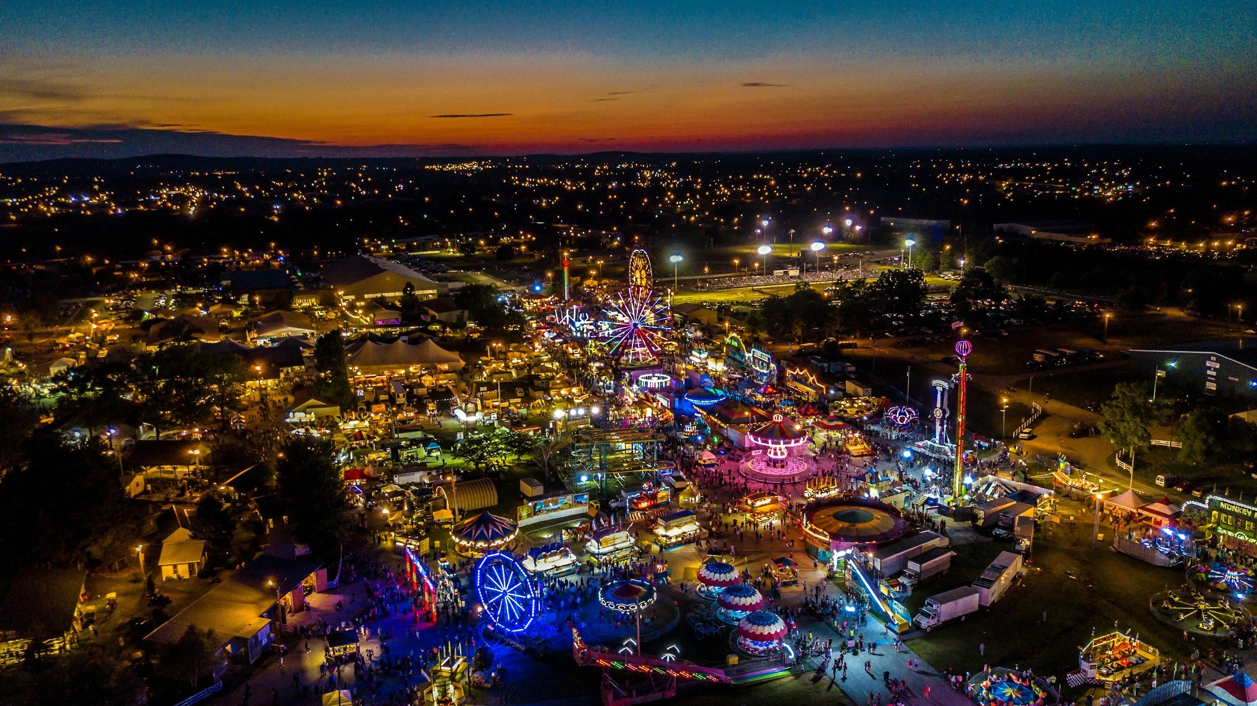 An aerial night view of a county fair with illuminated rides, tents, and stalls, surrounded by city lights and a sunset sky.