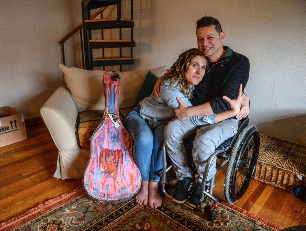 A man in a wheelchair hugging a woman who is sitting on his lap inside a living room. The woman is hugging a colorful guitar leaning against a sofa.