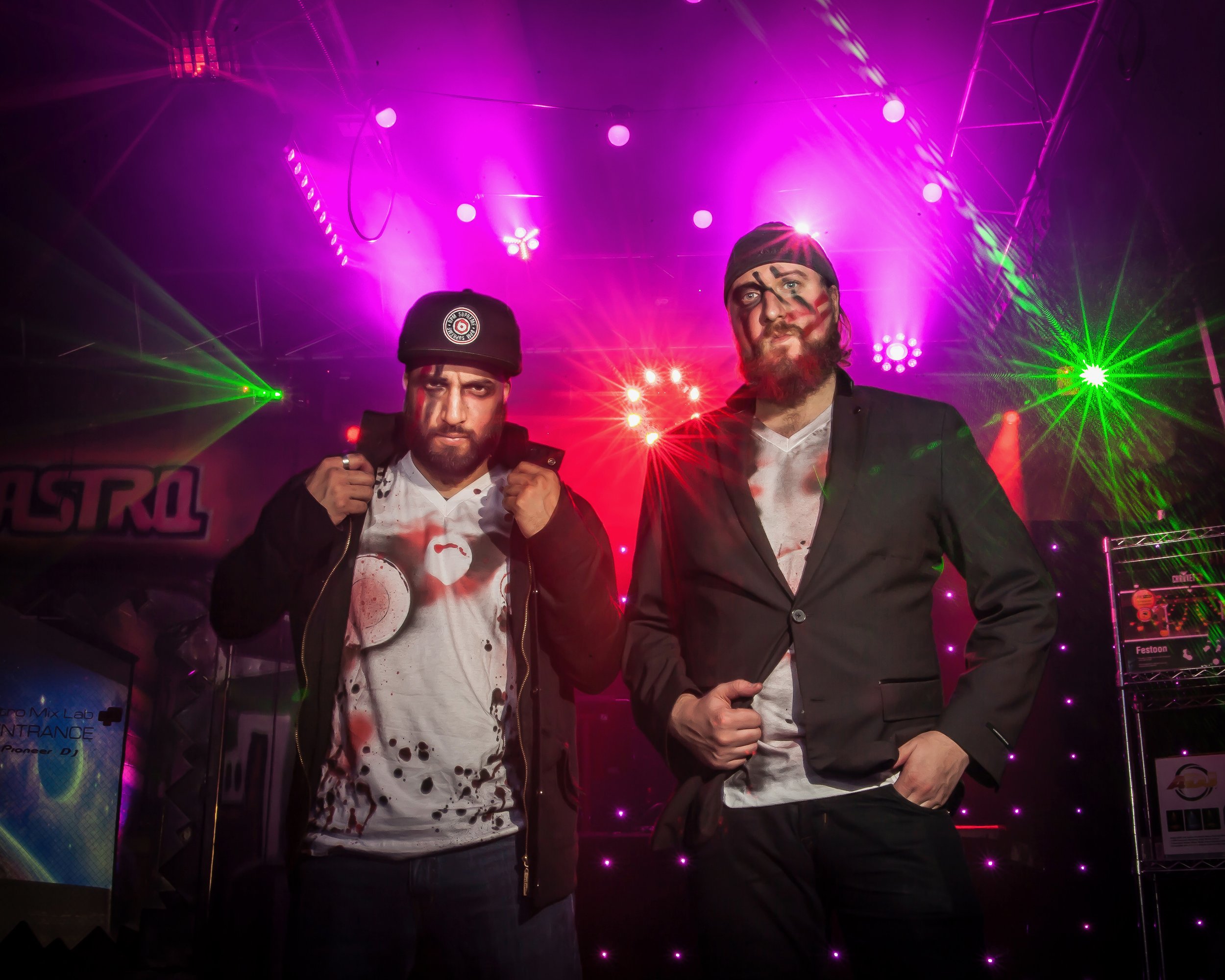 Two men with face paint standing in a colorful room with stage lights, wearing casual and formal attire, graffiti on the background wall.
