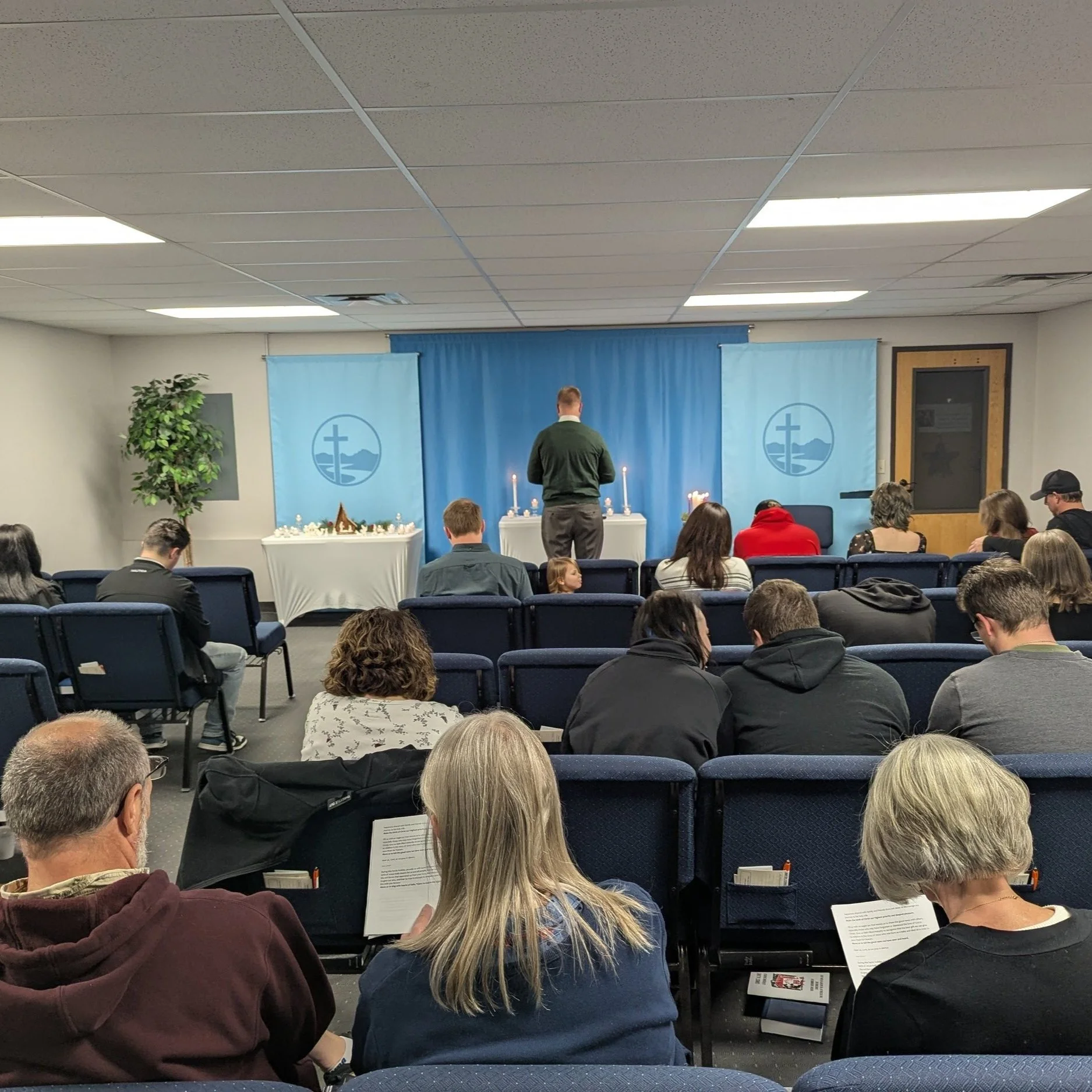 People sitting in chairs facing a small stage with a person standing in front of a table with candles in what appears to be a church or chapel setting.