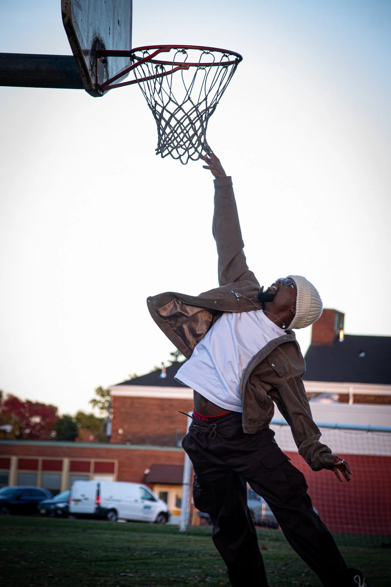 A man wearing sunglasses, a white beanie, a brown jacket, and black pants is reaching up to dunk a basketball into a hoop on an outdoor court during sunset.