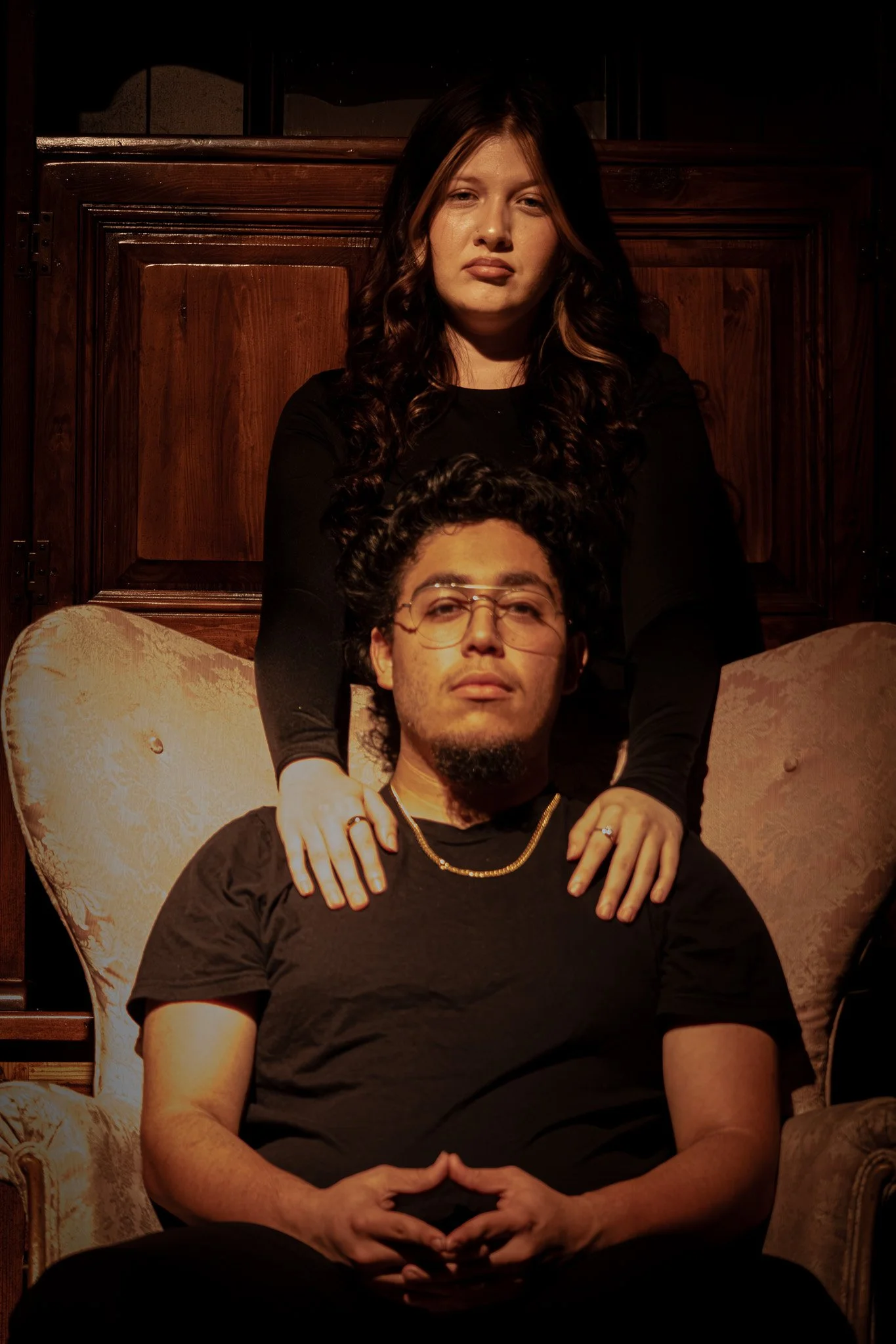 A man with glasses and a woman with long hair pose together in a dimly lit room with wooden furniture and beige chairs.