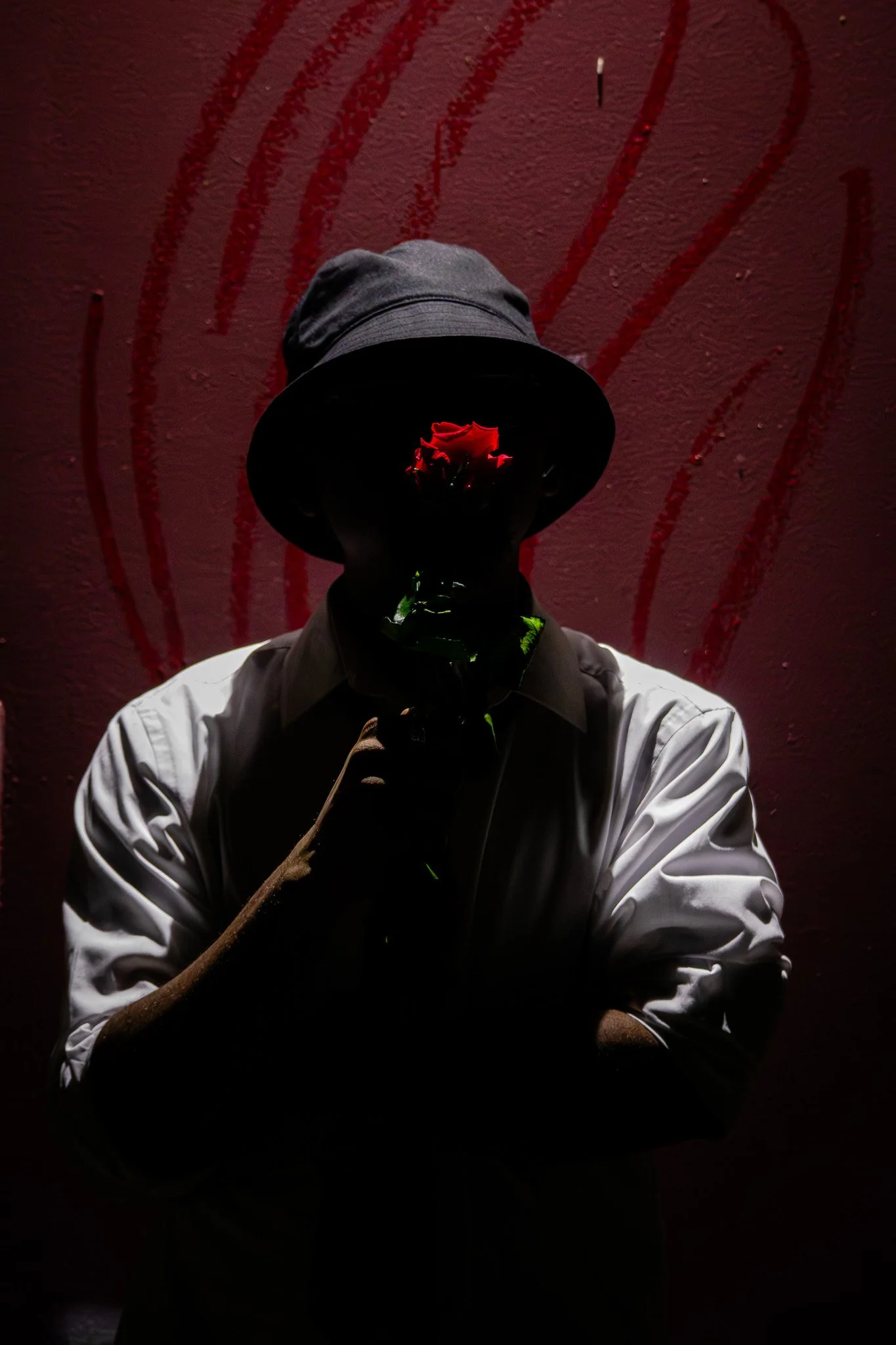 A person wearing a hat and white shirt holding a red rose, with their face obscured, standing in front of a red background with abstract red lines.