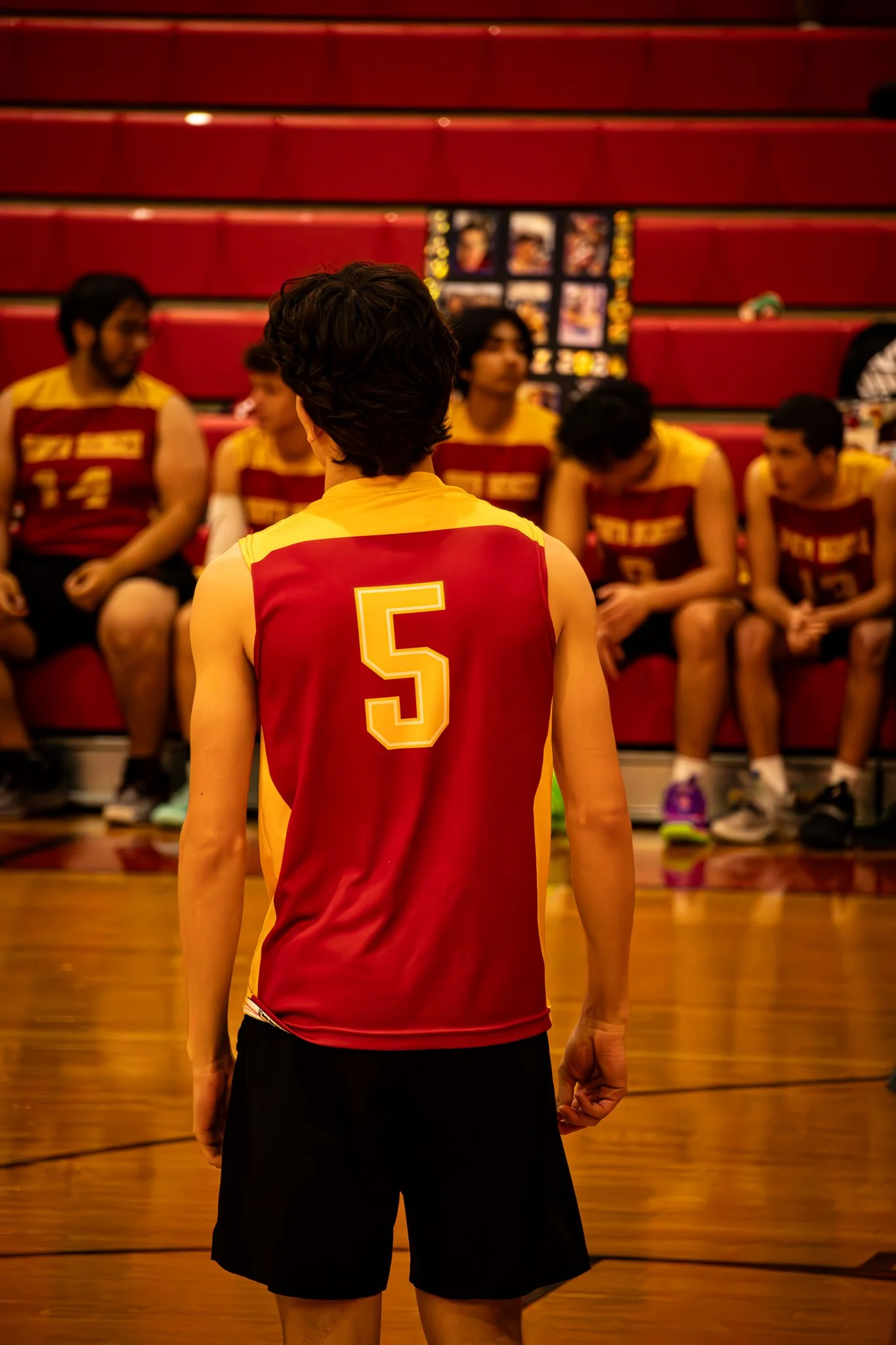 A volleyball player wearing a red and yellow jersey with the number 5 stands facing his teammates, who are sitting on the bleachers in the background.