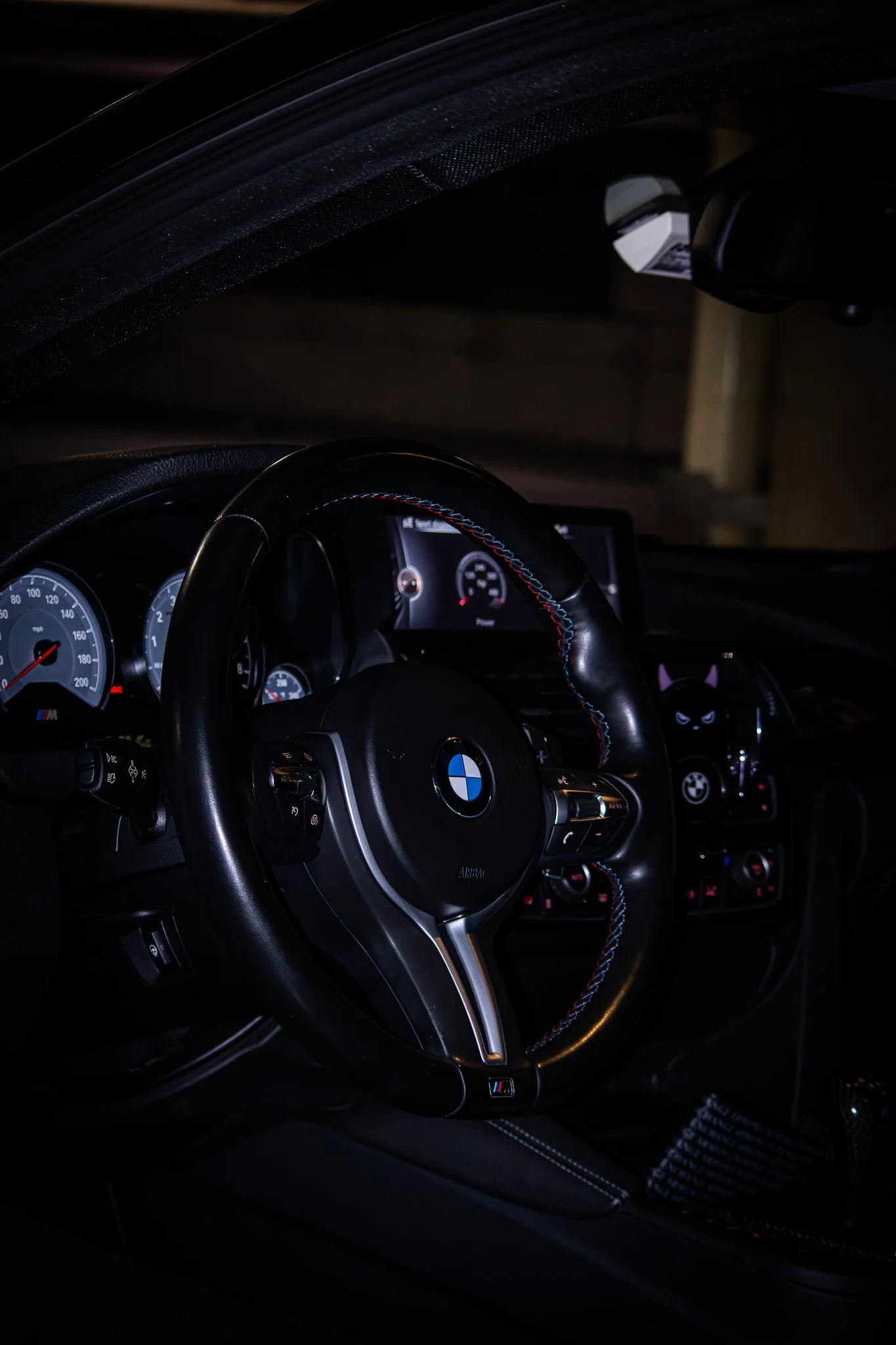 Interior of a BMW car showing the steering wheel with BMW logo, dashboard with speedometer and infotainment screen, and a gear shift with carbon fiber accents.