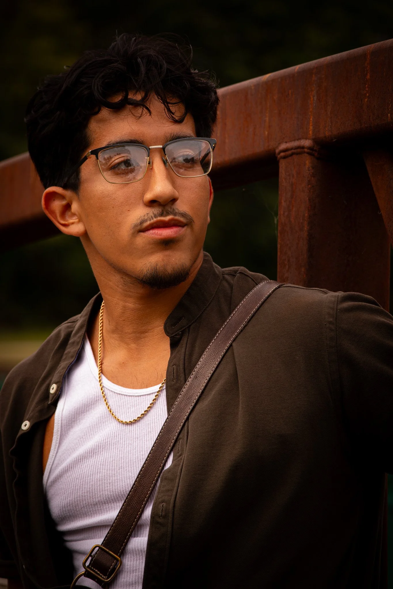 A young man with glasses, a goatee, and wearing a white tank top under a dark brown shirt, looking to the side outdoors next to a rusted metal structure.