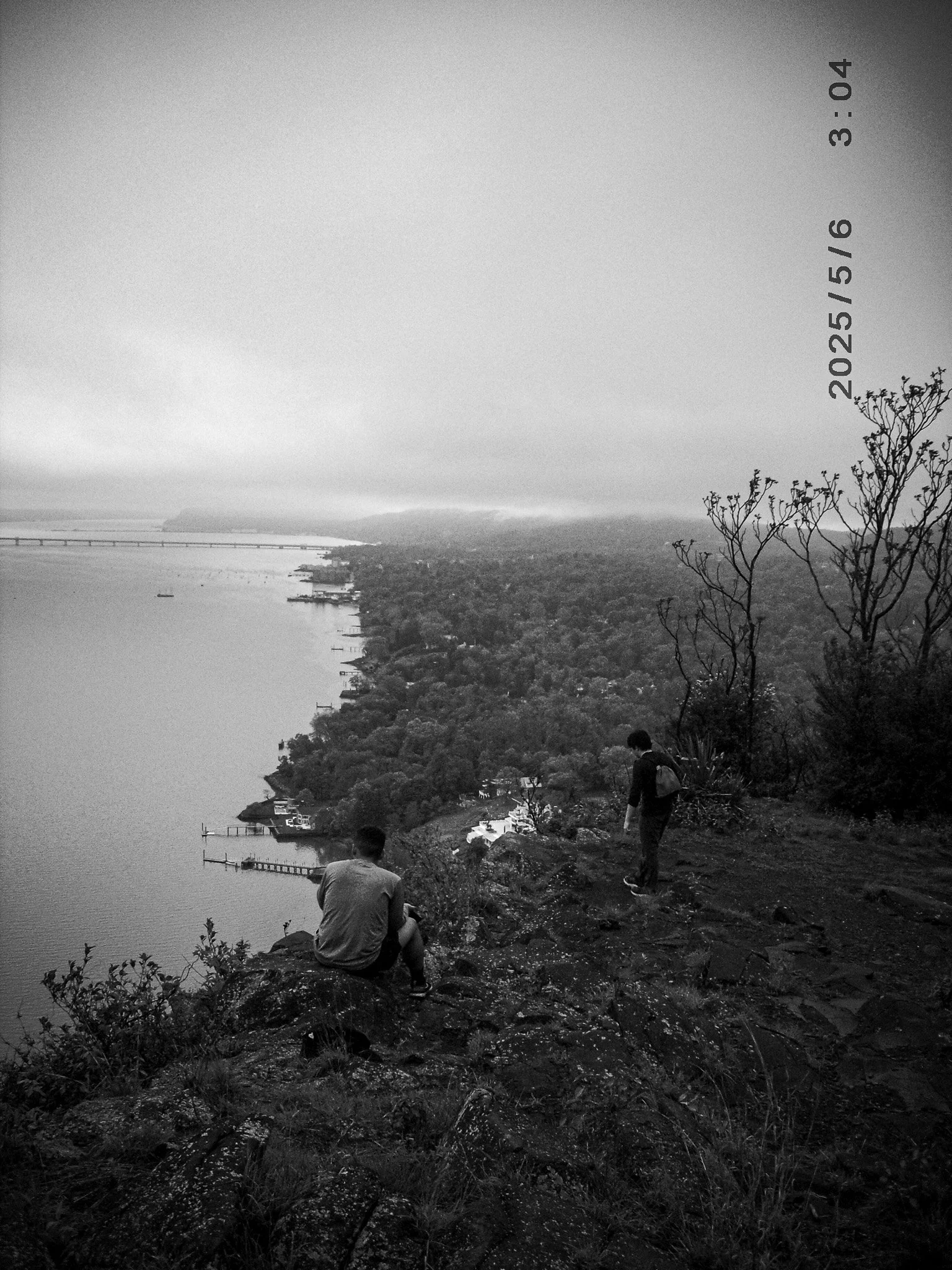 Two people hiking on a rocky trail overlooking a body of water, with a tree on the right and a distant bridge in the background, in black and white.