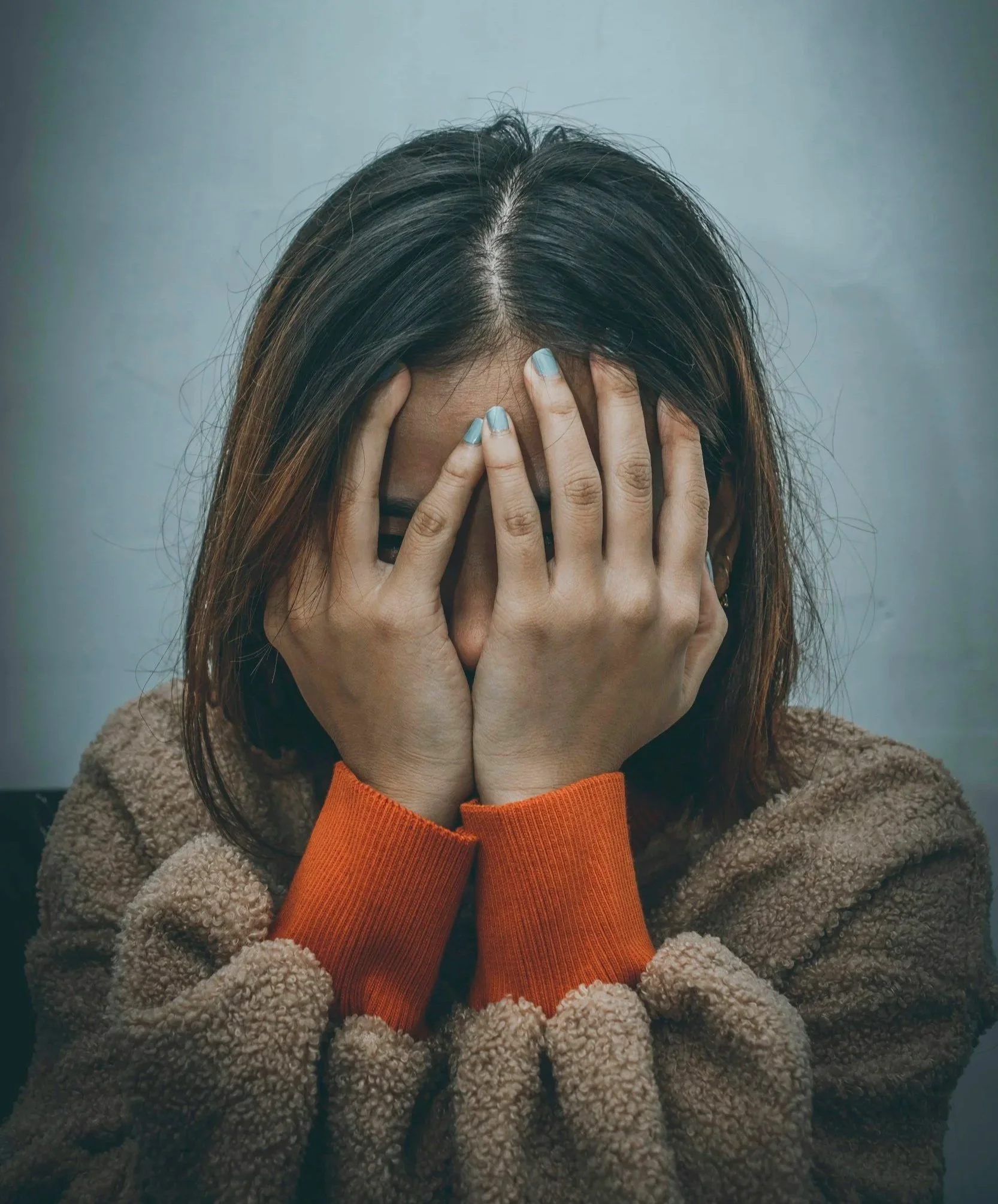 A woman covering her face with her hands, wearing a brown fleece jacket with orange cuffs.