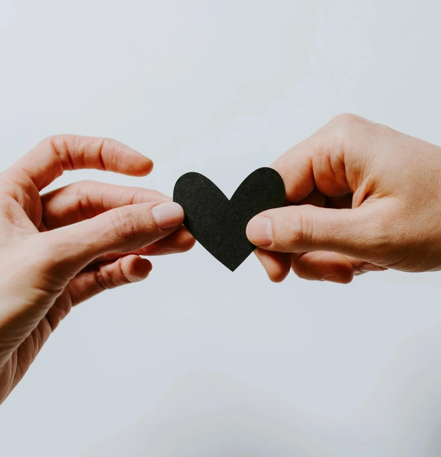Two hands exchanging a black paper heart against a plain light gray background.