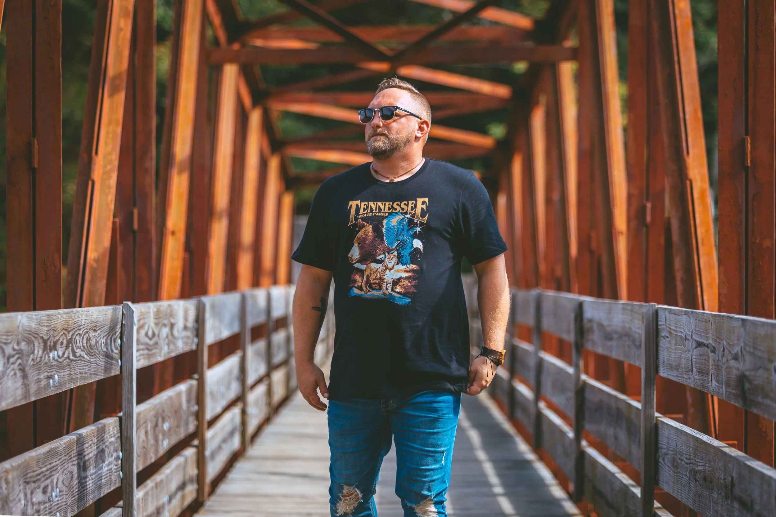 A man walking on a wooden bridge outdoors, wearing sunglasses, a black Tennessee State Parks T-shirt, and ripped jeans.