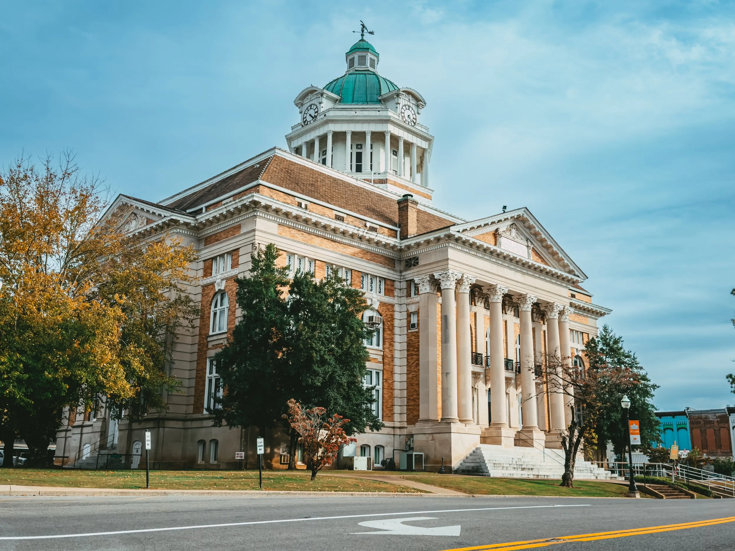 A historic courthouse building with a clock tower, stone steps, large columns at the entrance, and trees in the foreground under a partly cloudy sky.