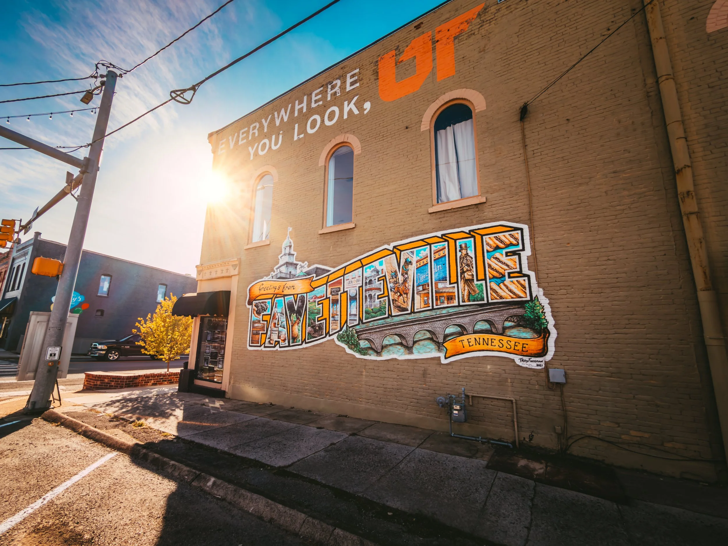 Colorful mural on a brick building in Tennessee that reads "Greetings from Tennessee" with illustrations of landmarks and local scenery, under a clear sky with sunlight.