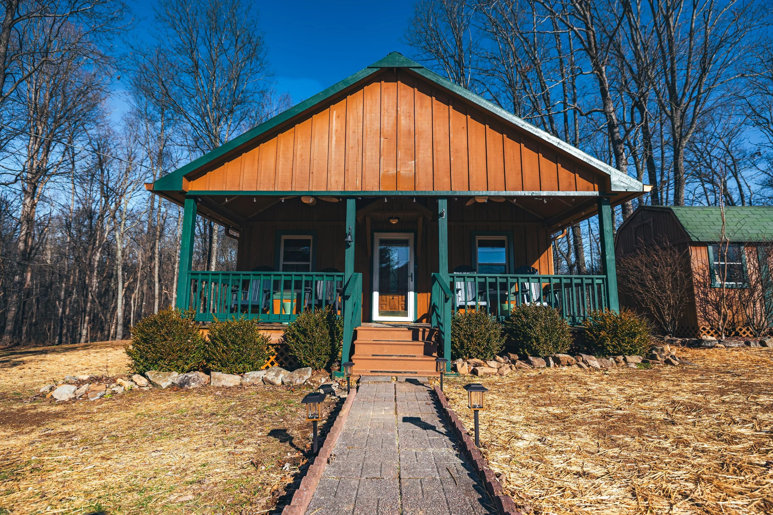 Front view of a wooden house with a covered porch, green railings, steps leading up to the front door, surrounded by bushes, with a pathway and outdoor lights in front, and trees in the background.