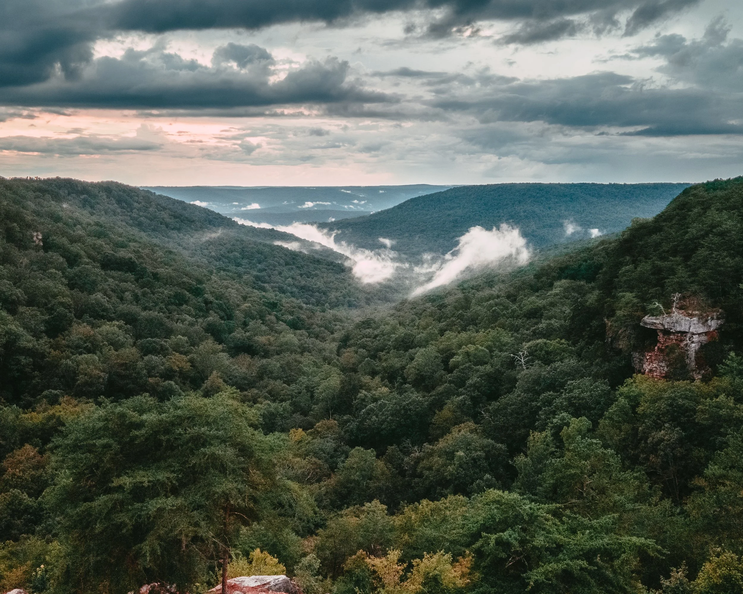 View of a valley with lush green forests, rolling hills, and distant mountains under a cloudy sky with patches of sunlight and mist in the air.