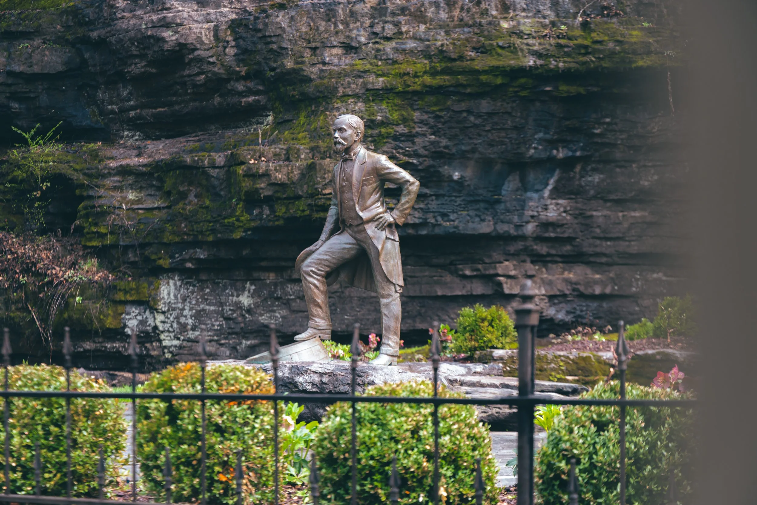 Bronze statue of a man with a beard in a suit, standing with one hand on his hip and one foot on a rock, outdoors with a rocky background and small bushes in front, enclosed by a black metal fence.