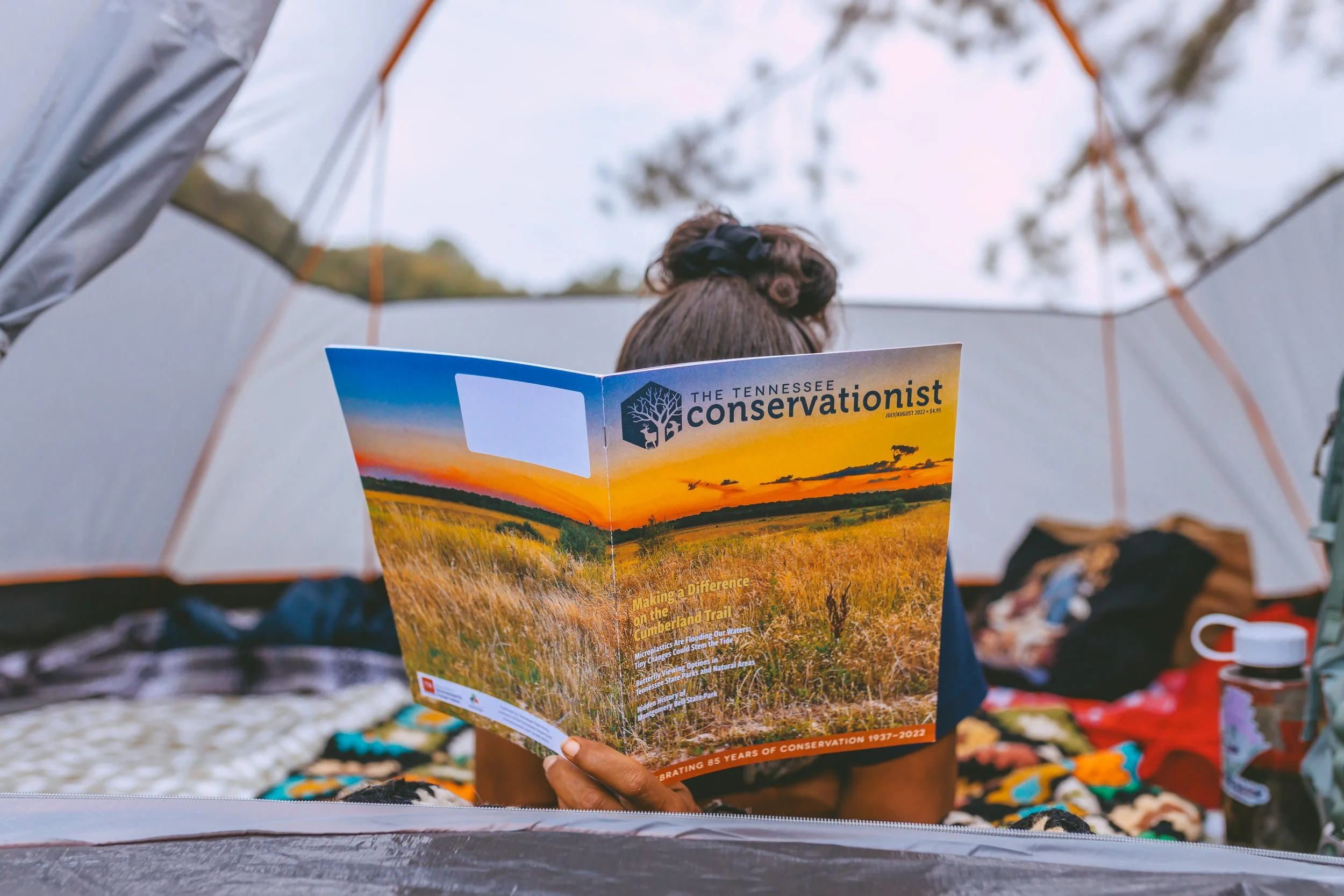 Person sitting in a tent reading a magazine titled "The Tennessee Conservation" with a landscape of grass and trees at sunset on the cover.