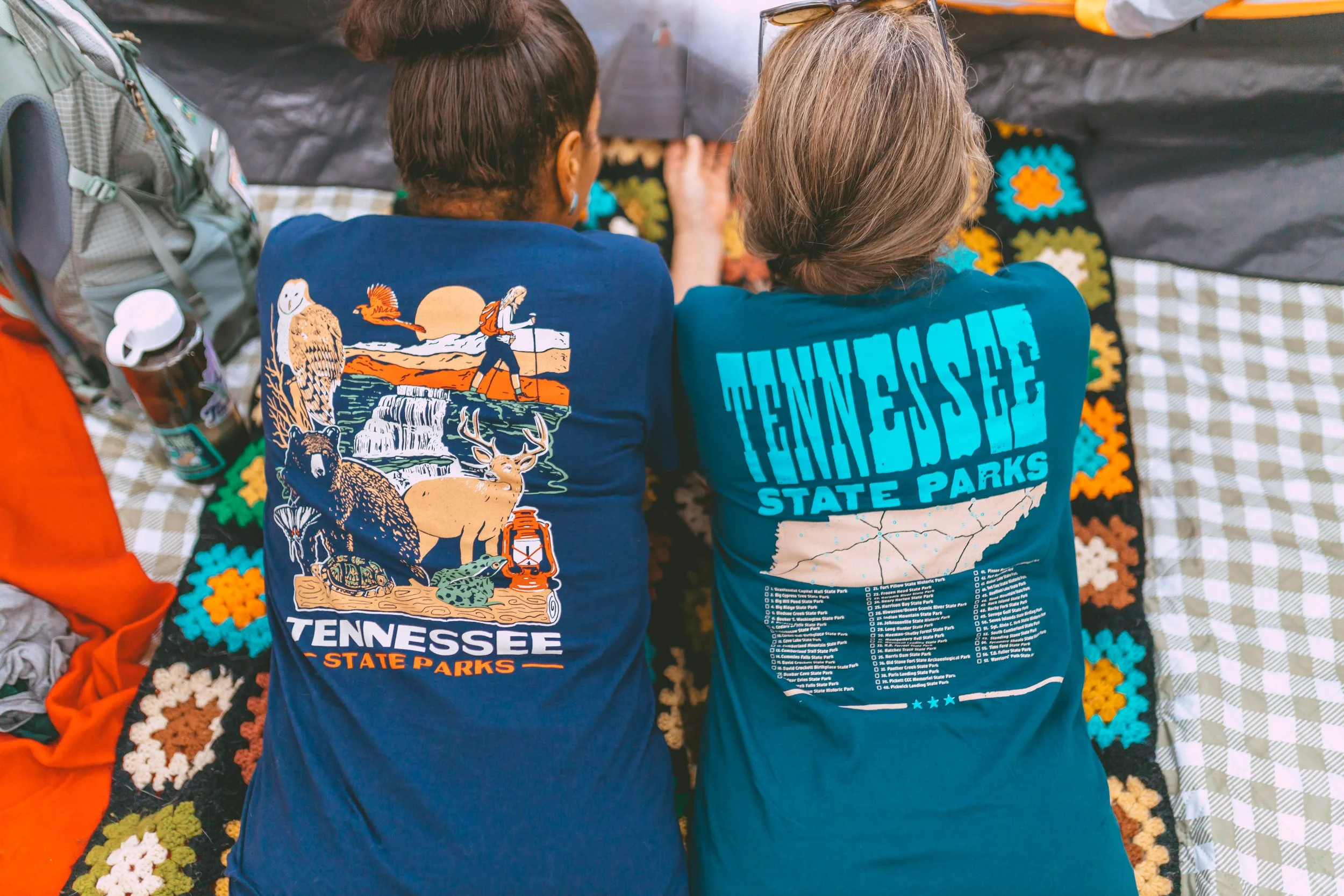 Two people sitting on a colorful afghan blanket with their backs to the camera, wearing Tennessee State Parks shirts at an outdoor setting with camping gear nearby.