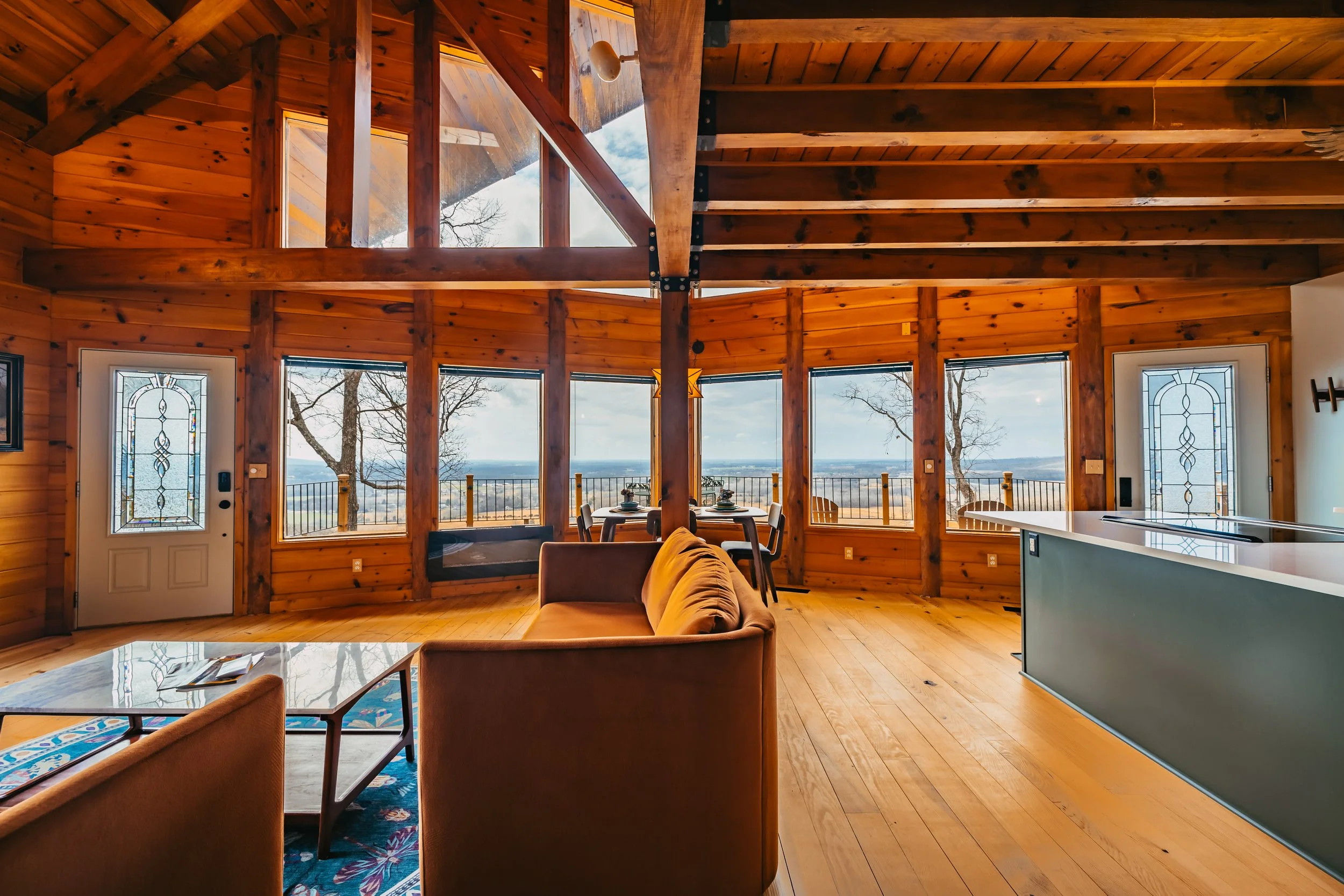 Interior of a wooden house with large windows overlooking a landscape. Contains a sofa, coffee table, dining table with chairs, and a kitchen island.