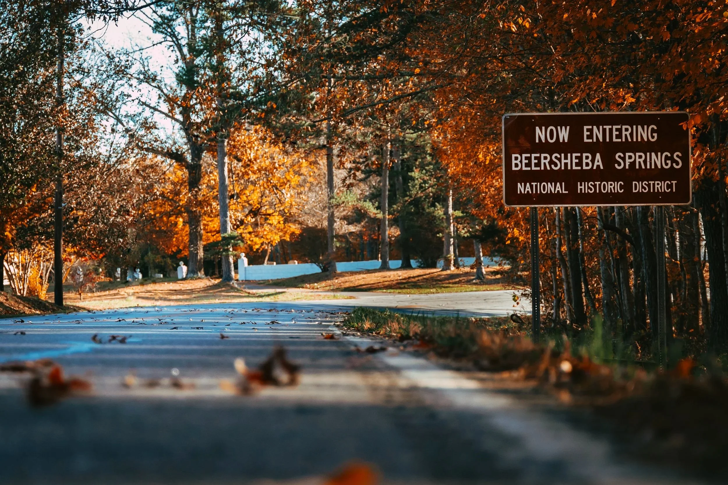 A road leading into Beersheba Springs, a historic district, with fall foliage and trees on both sides. A brown sign on the right reads: 'Now Entering Beersheba Springs National Historic District.'