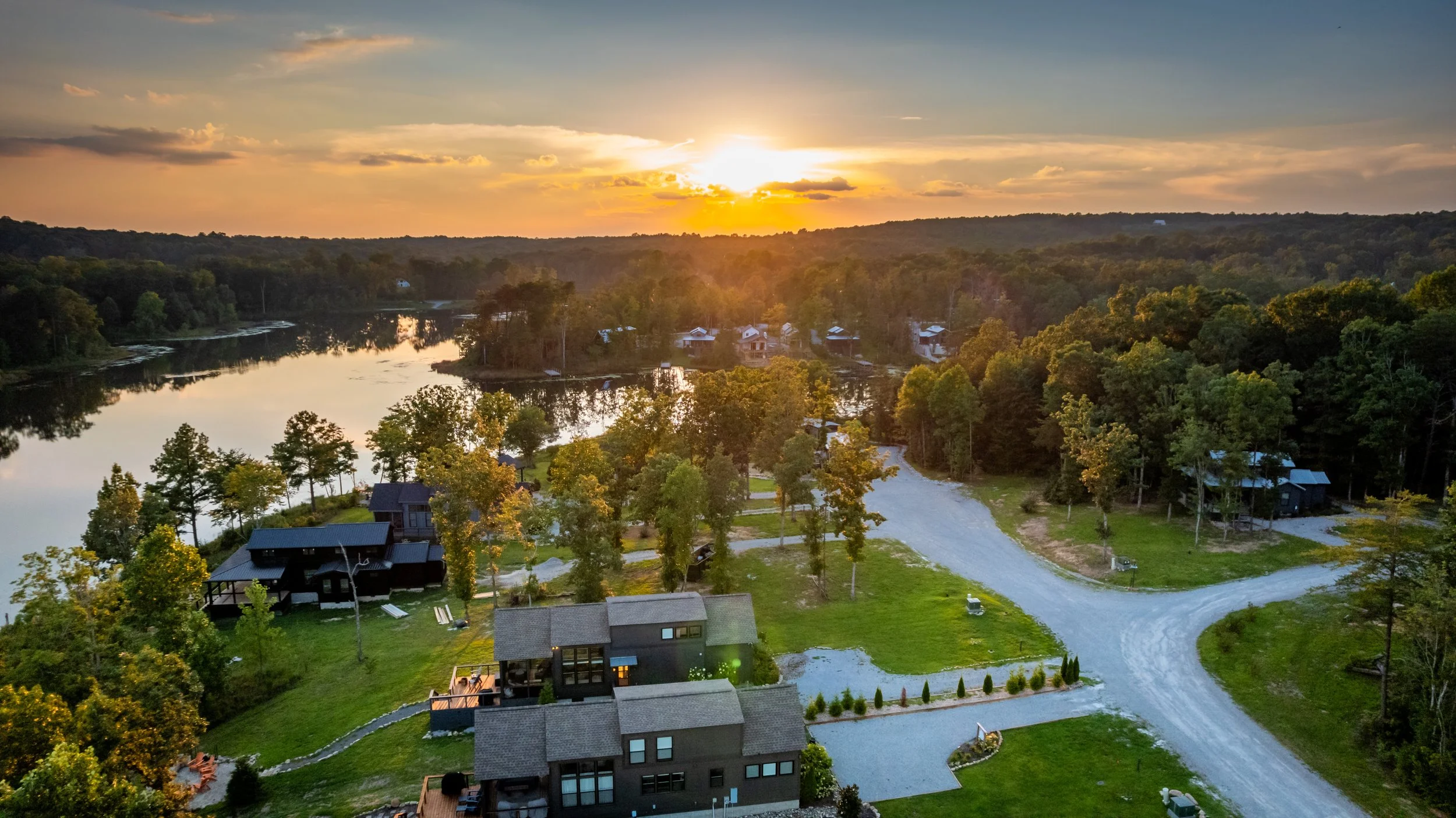 Aerial view of a lakefront community at sunset with houses, green lawns, and trees along a winding gravel road.