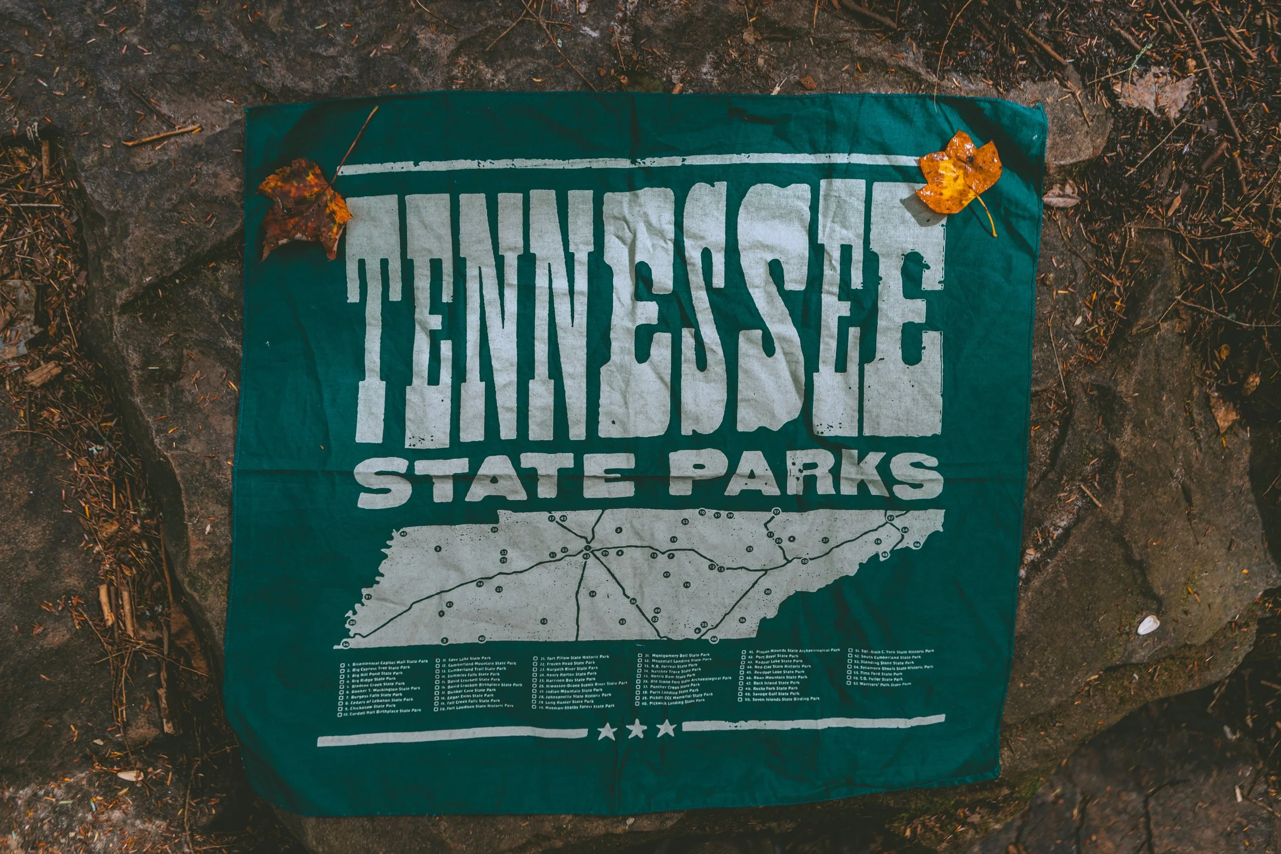 Tennessee State Parks map and logo on a green cloth, with fallen autumn leaves on top, placed on the ground.
