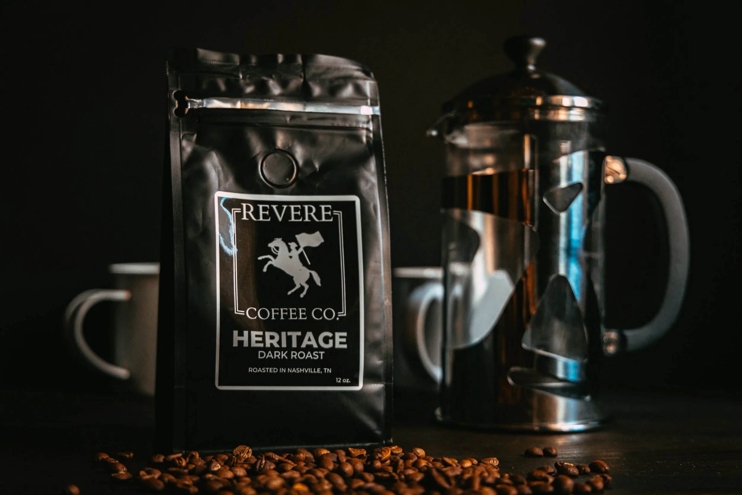 A bag of Revere Coffee Co. Heritage Dark Roast coffee beans, a moka pot, and a white coffee mug on a dark surface with scattered coffee beans.