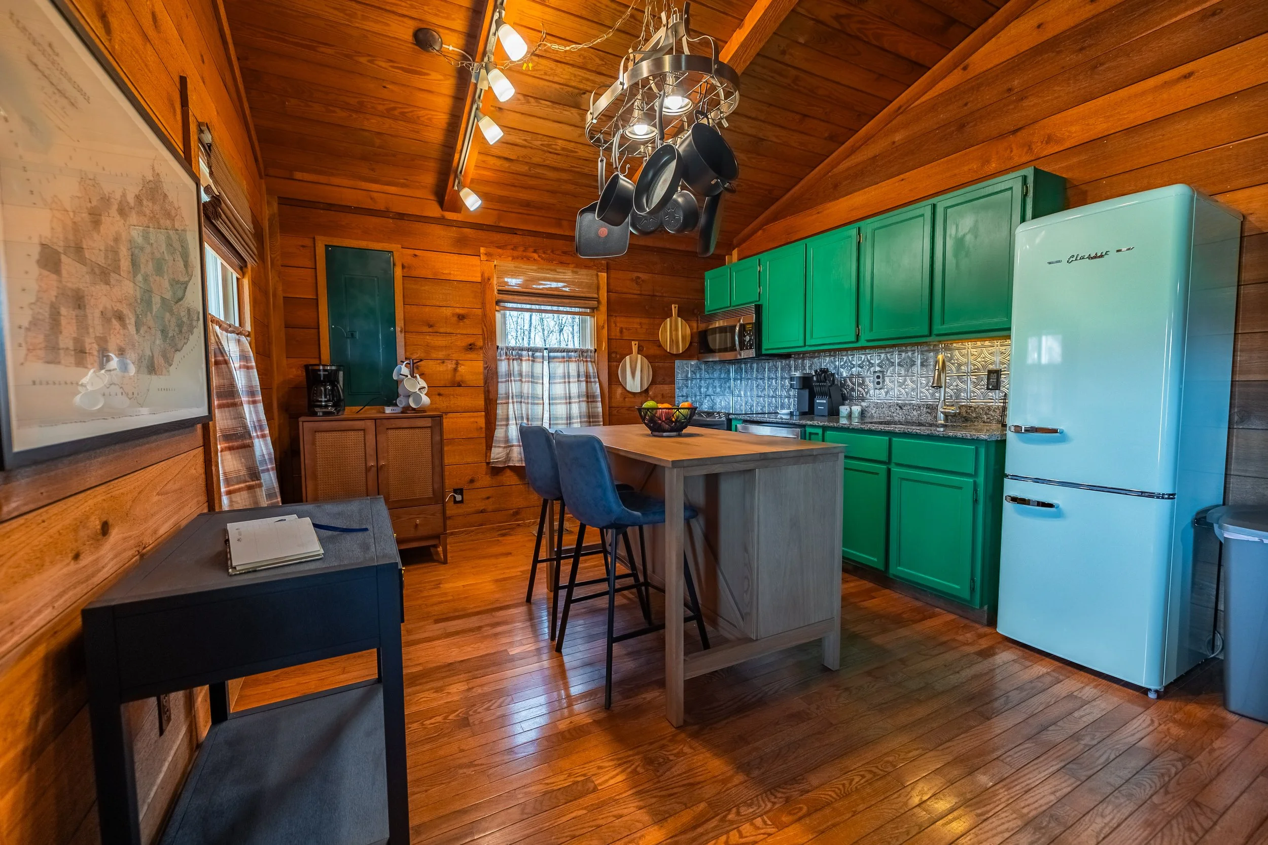 A cozy rustic kitchen with wooden walls and floors, green cabinets, a retro refrigerator, a wooden kitchen island with bar stools, and a pot rack hanging from the ceiling.