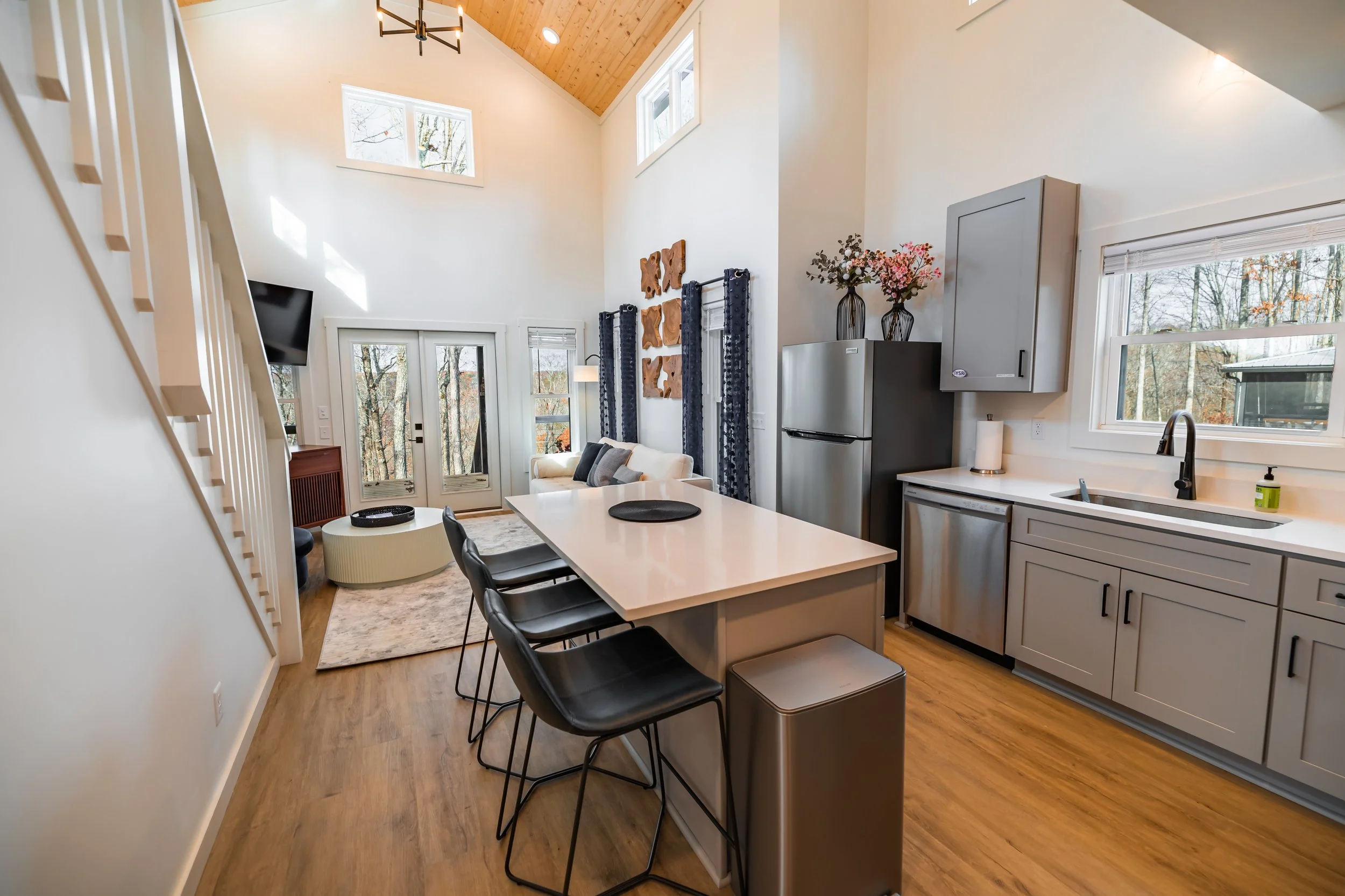 Open-concept living room and kitchen with high vaulted ceiling, hardwood floors, and large windows showing trees outside, featuring a dining table, black bar stools, kitchen cabinets, a refrigerator, and a white sofa.
