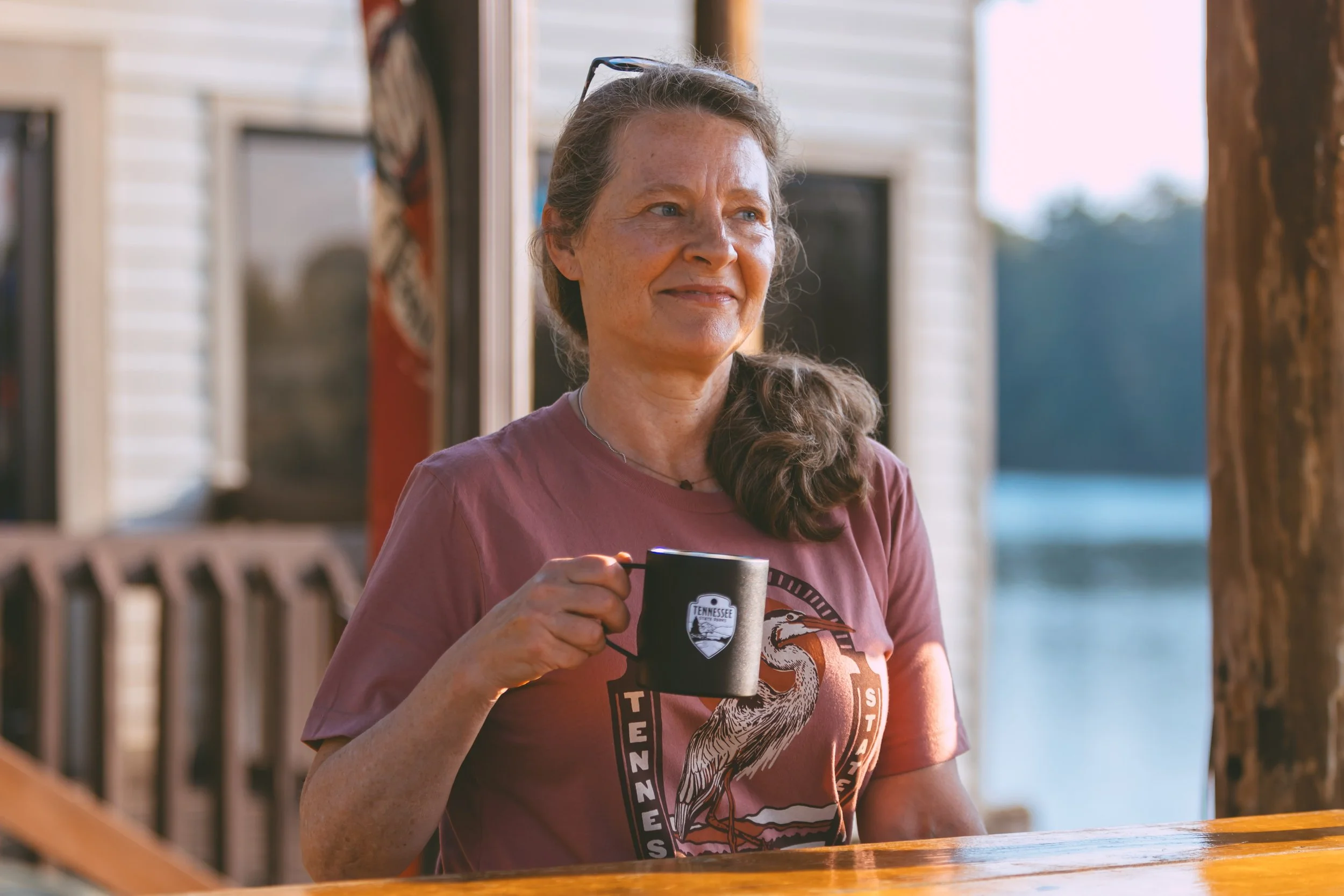 A woman with long brown hair, wearing a Tennessee-themed t-shirt, is holding a black mug with a Tennessee emblem on it. She is sitting outdoors near a body of water and a wooden post, with a house in the background, on a sunny day.