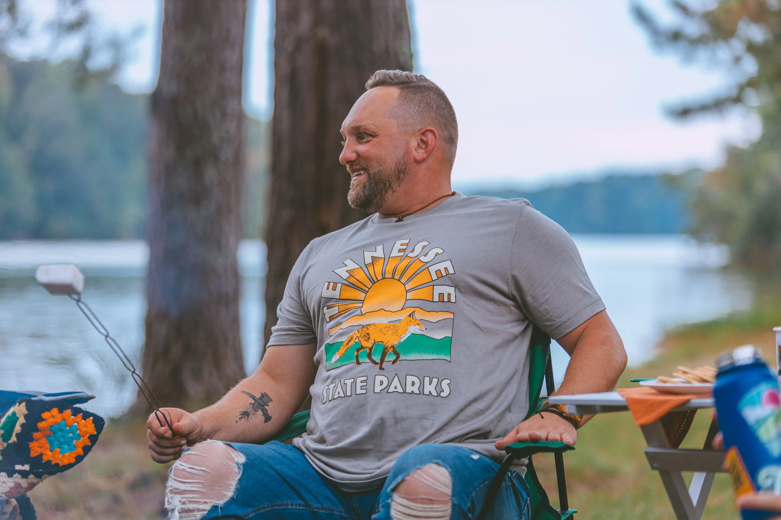 A smiling man sitting outdoors near a lake, wearing a gray T-shirt with a sunset and wolf design that reads "ESEE State Parks," holding a marshmallow roasting stick, with a table of snacks nearby.