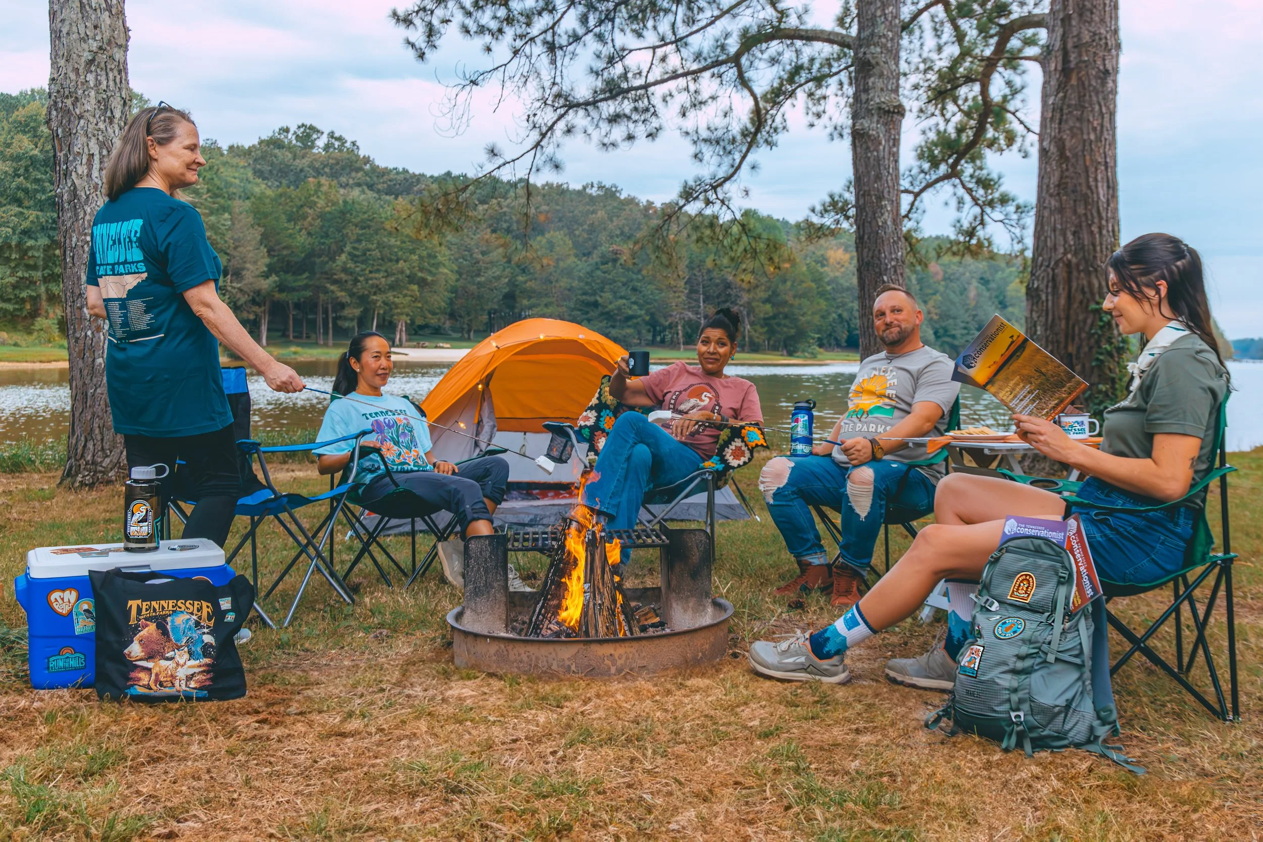 Group of people camping outdoors by a lake with a campfire, chairs, and a yellow tent, surrounded by trees.