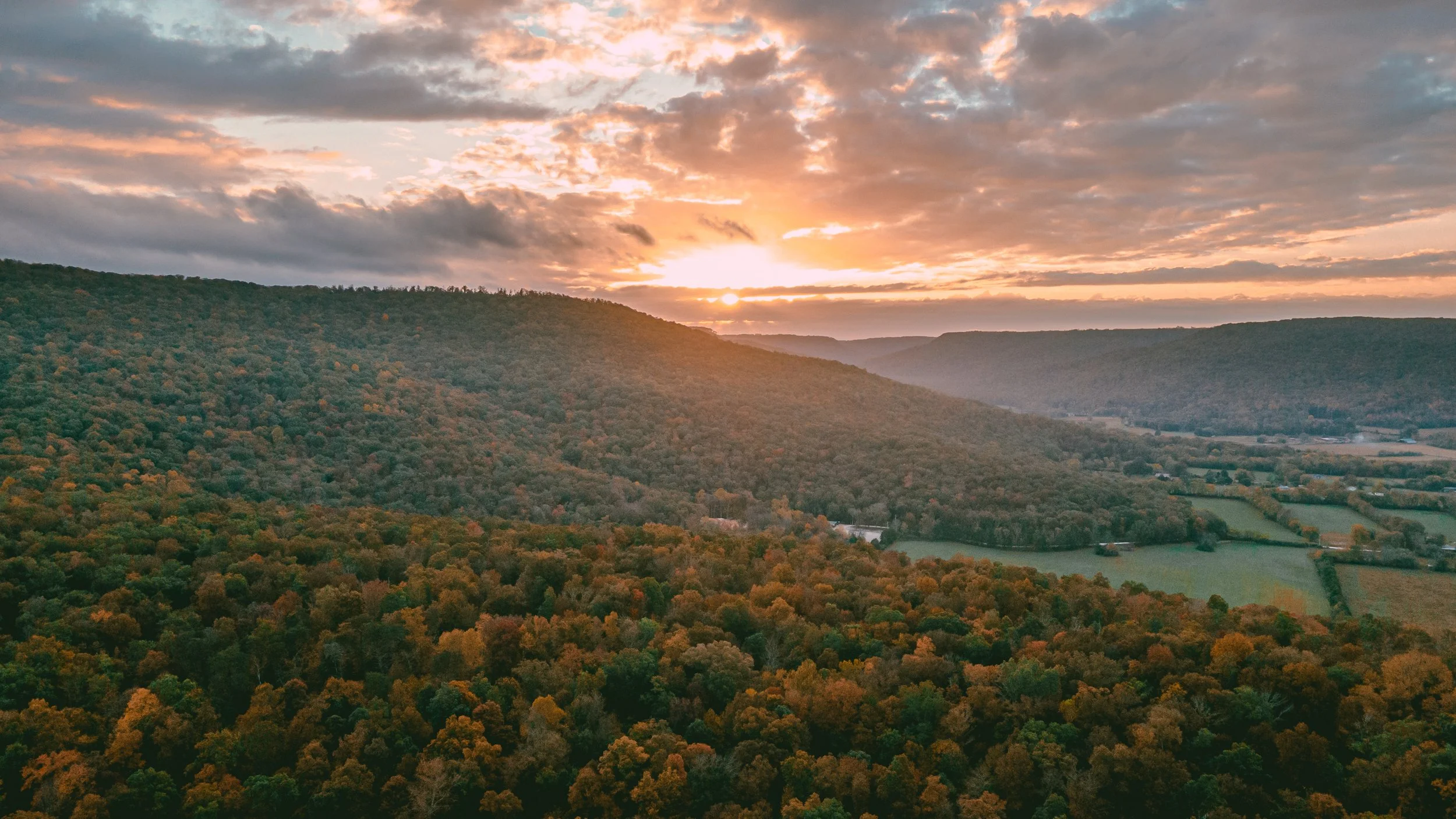 A scenic view of rolling hills covered in autumn foliage under a partly cloudy sky at sunset.