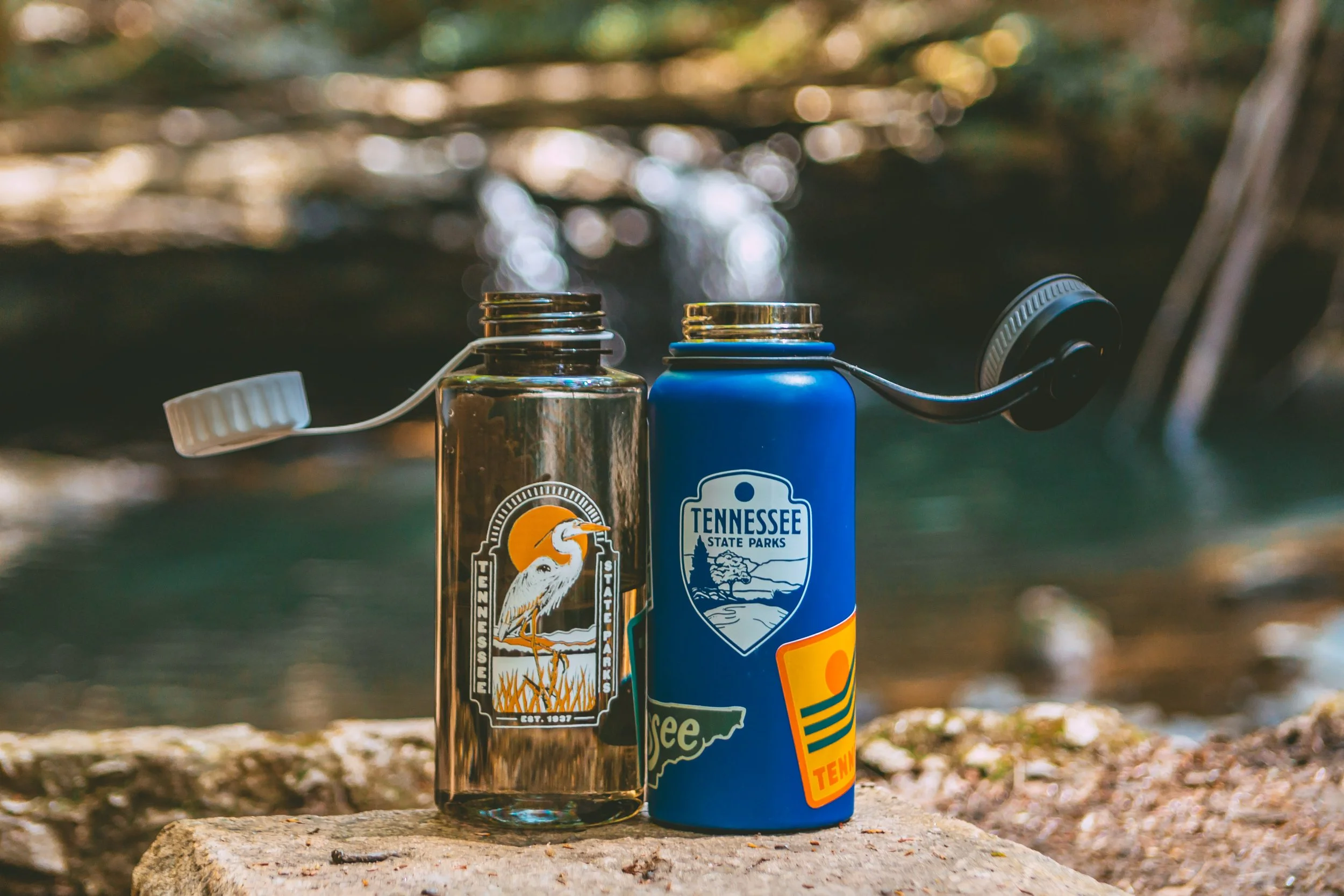 Two flasks, one metallic with a Tennessee State Parks logo featuring a heron, and the other blue with a Tennessee State Parks emblem and colorful design, are placed on a rock near a stream in a wooded area.