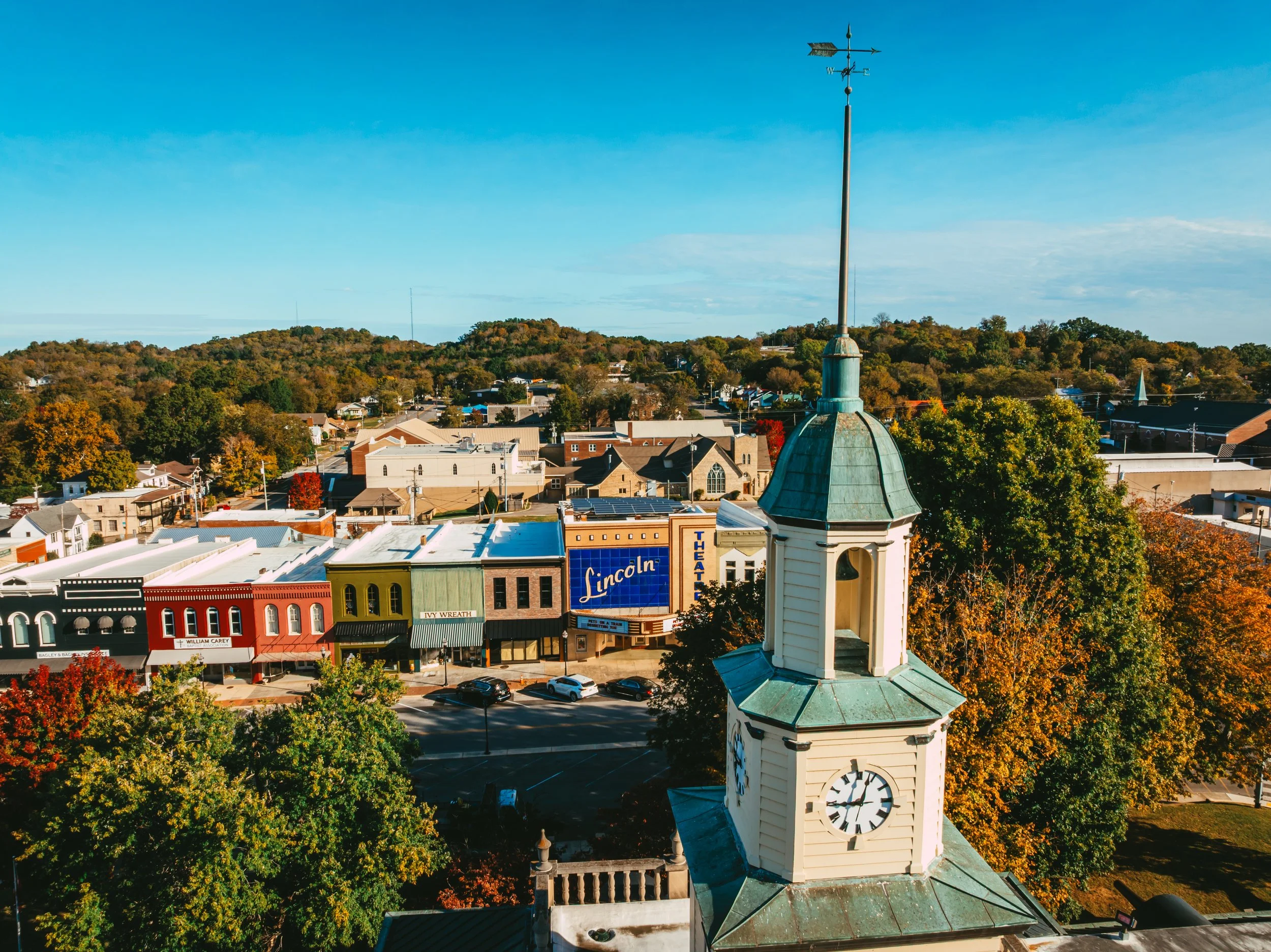 Aerial view of a small town with colorful buildings, a church steeple with a clock, and trees with autumn foliage, against a backdrop of hills and a blue sky.