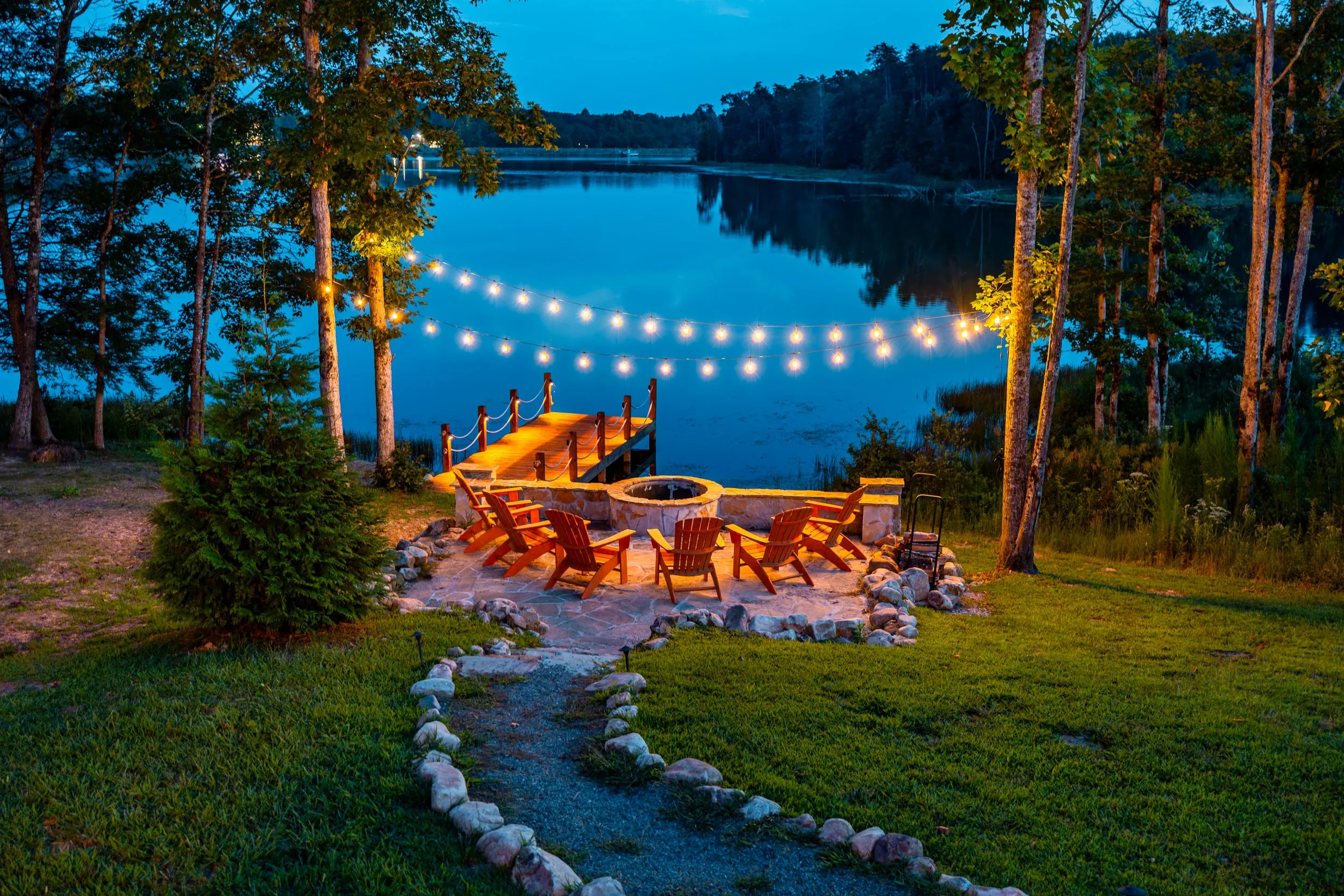 A lakeside outdoor patio with Adirondack chairs surrounding a fire pit, a small dock extending into the water, string lights hanging overhead, and trees on a calm evening.