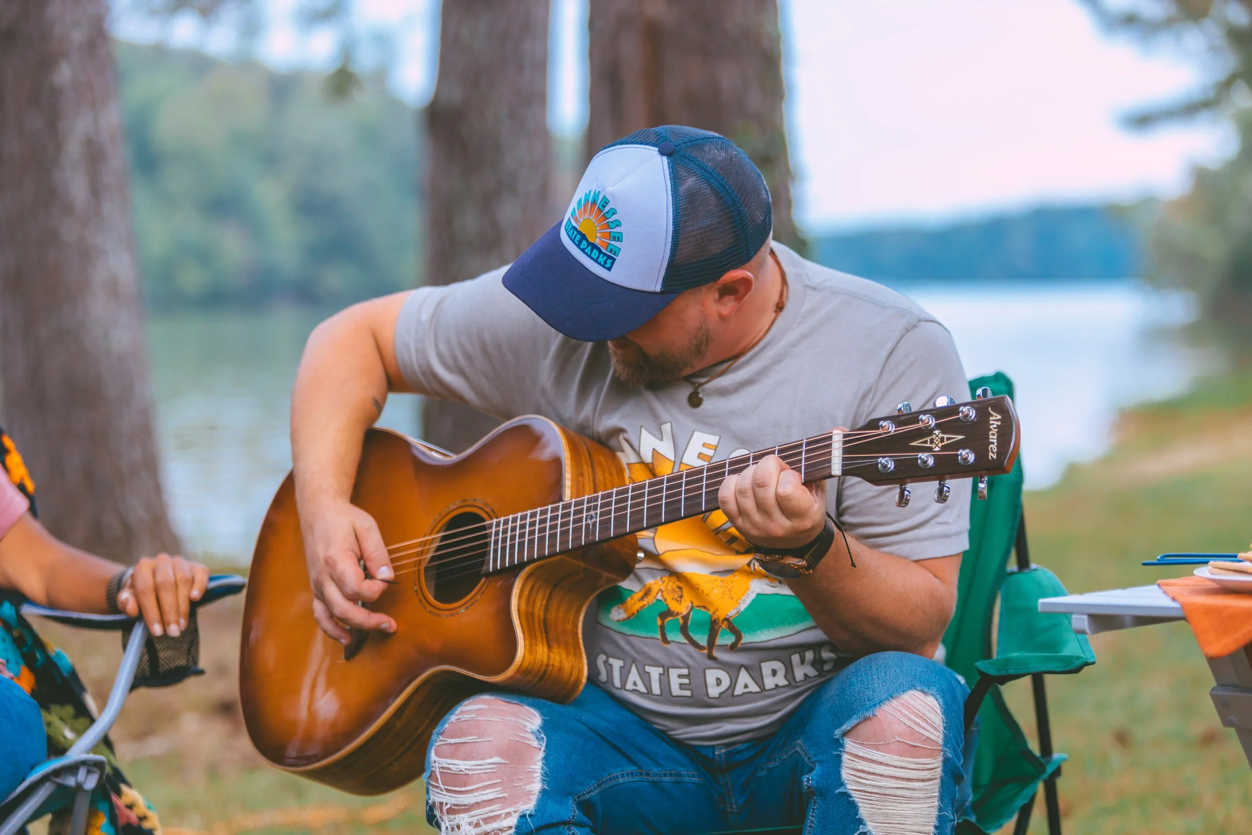 Man sitting outdoors playing an acoustic guitar near trees and a body of water, wearing a blue cap, gray T-shirt, and ripped jeans.