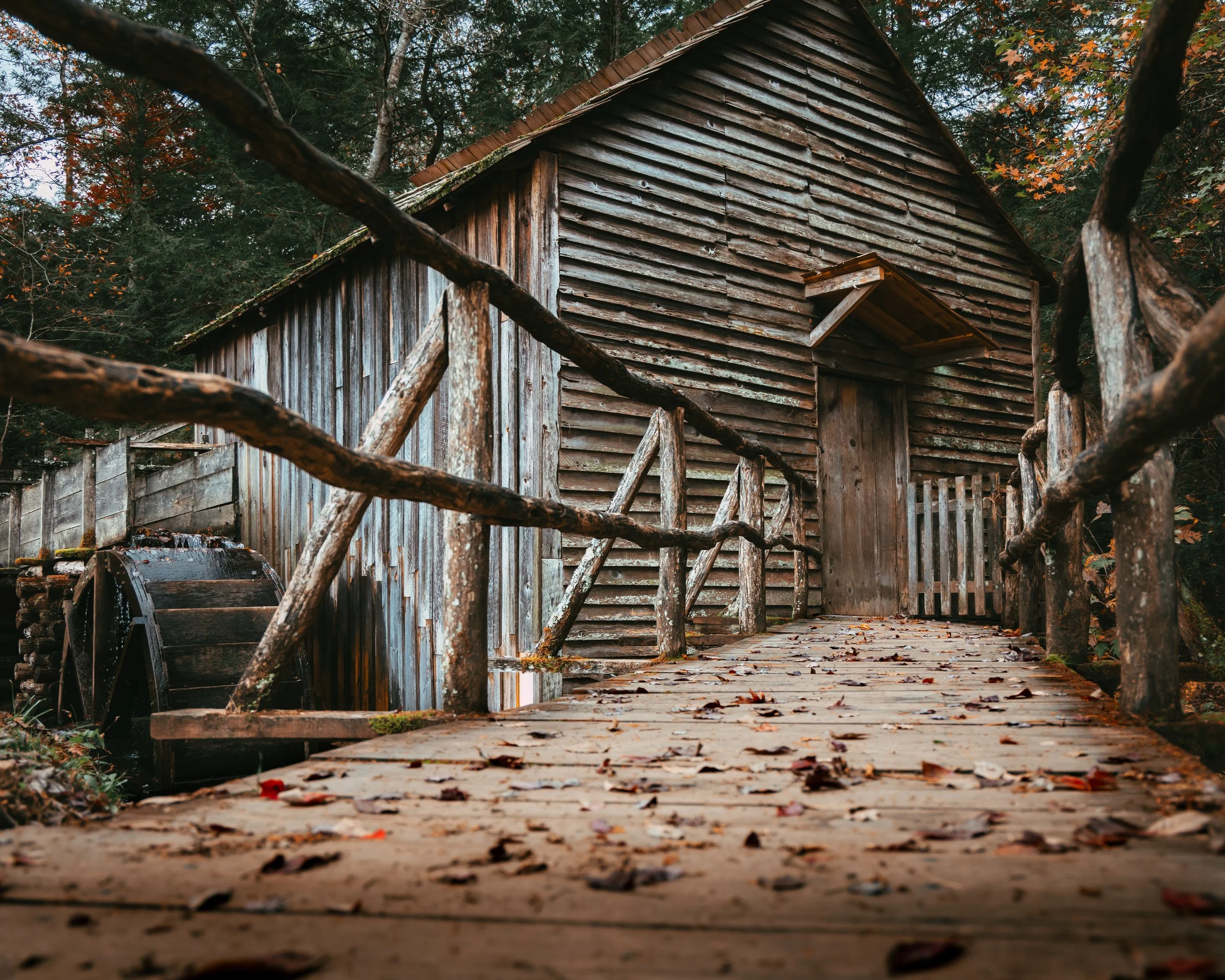 A rustic wooden walking bridge leading to a weathered barn surrounded by autumn trees.