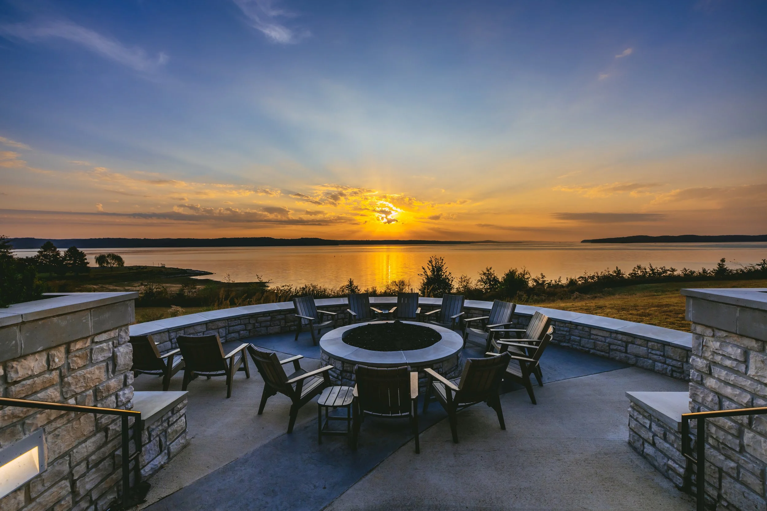 Sunset over a lake viewed from a stone patio with a circular fire pit and eight wooden chairs arranged around it.