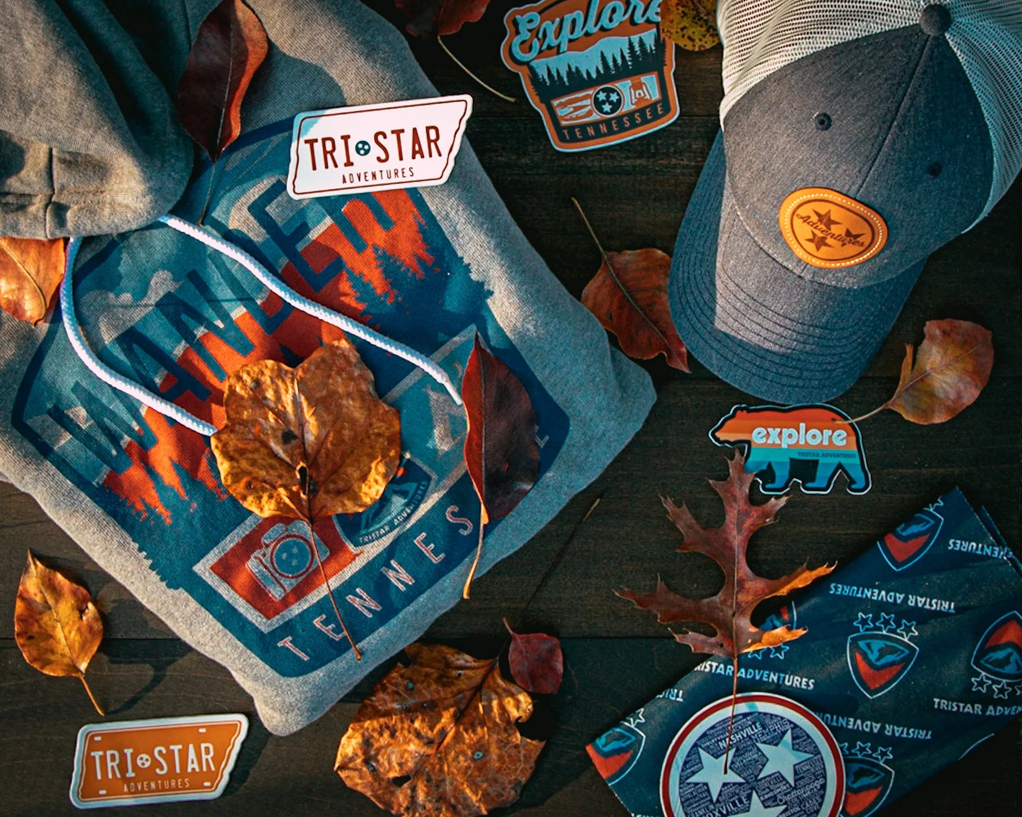 An outdoor display with Tennessee-themed clothing and accessories, including a gray hoodie with a mountain and tree design, a gray cap with a leather patch, and a bandana with Tennessee symbols, surrounded by fallen autumn leaves.