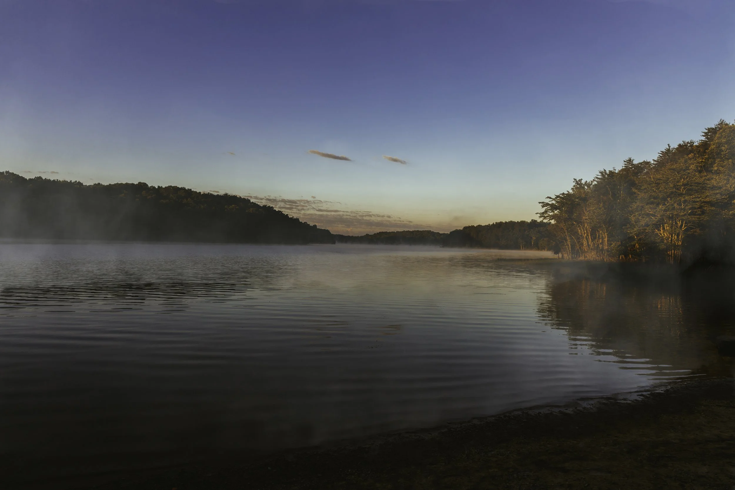A calm lake at sunrise with some mist rising, surrounded by trees on both sides, under a clear blue sky.