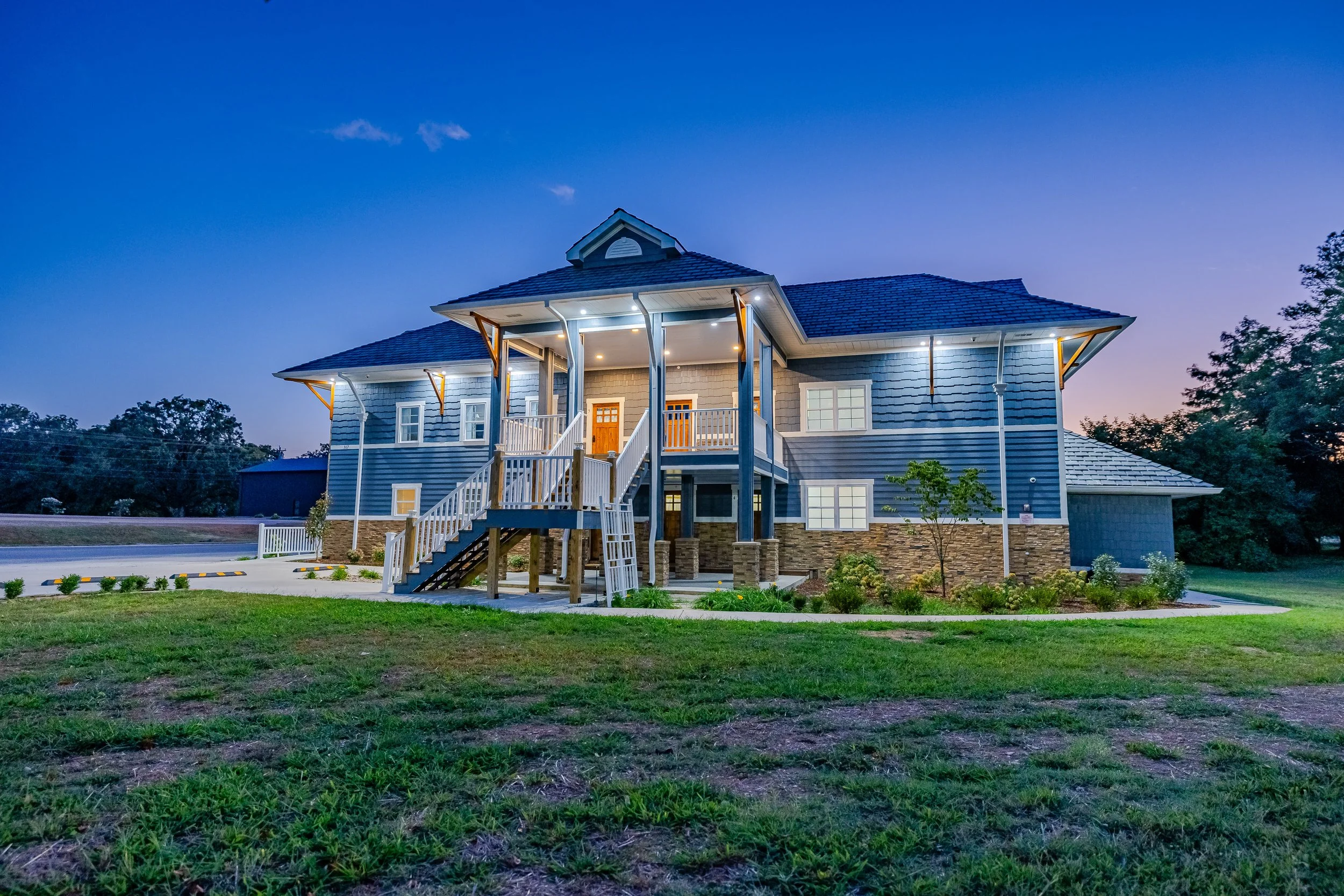 A two-story blue house with a front porch and stairs, surrounded by green grass and trees at dusk.