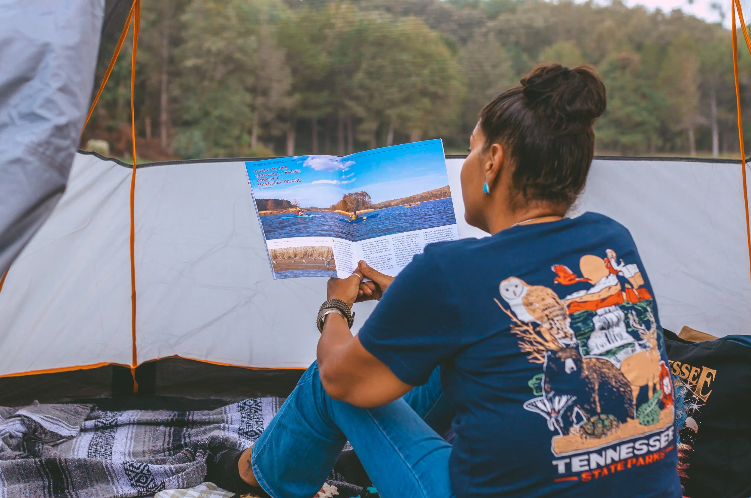 Person sitting in a tent reading a magazine with a lake and trees visible outside.