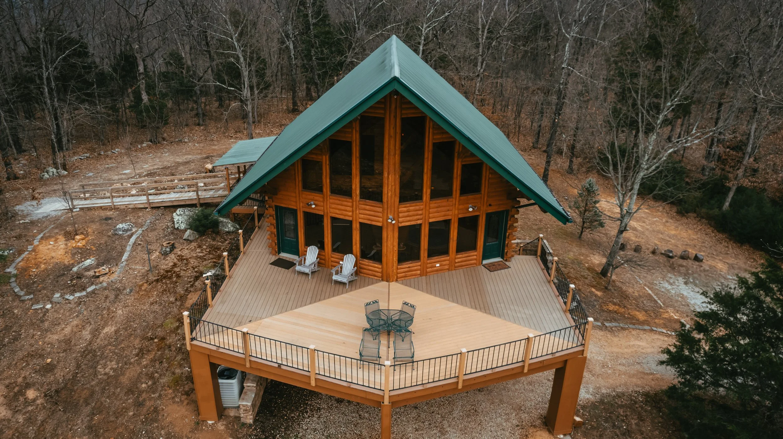 Aerial view of a wooden cabin with a green gabled roof, large glass windows, and a spacious elevated deck with outdoor furniture, surrounded by a forest with leafless trees.