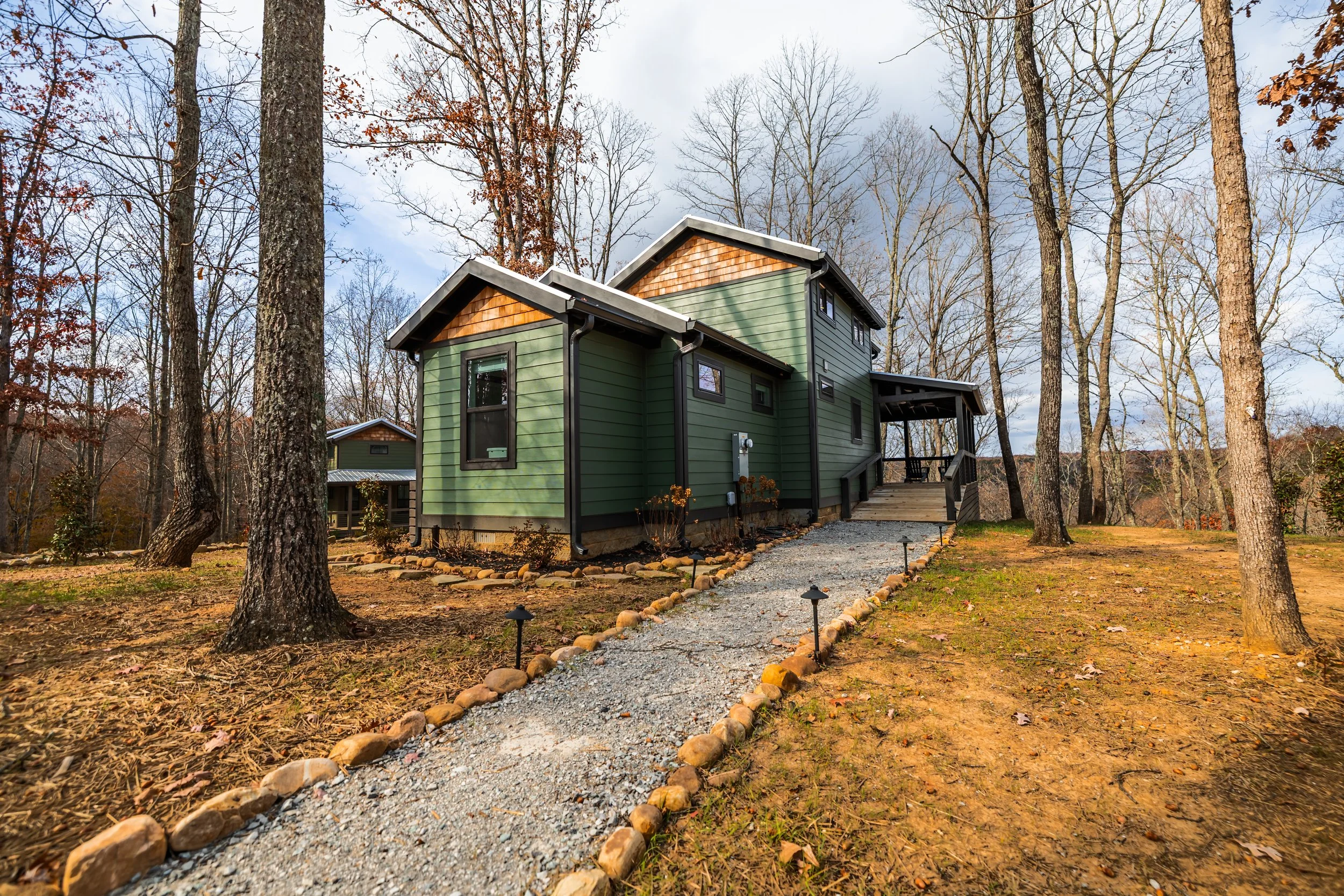 A green house with black trim and a small porch located in a wooded area during late fall or winter.