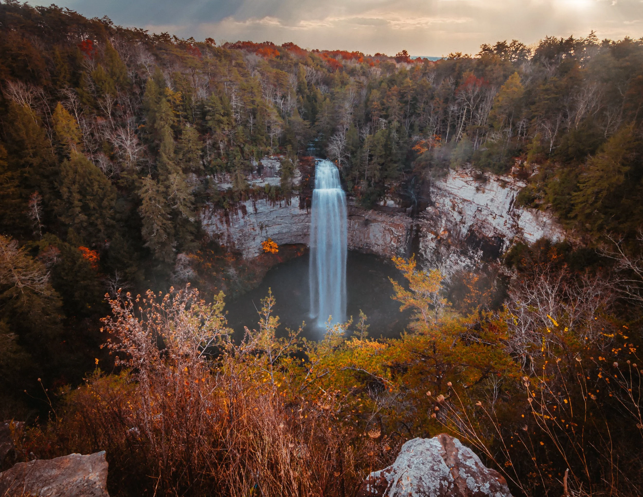 Aerial view of a waterfall in a forested area during autumn with orange, red, and green foliage surrounding a water cascade flowing into a pool below.