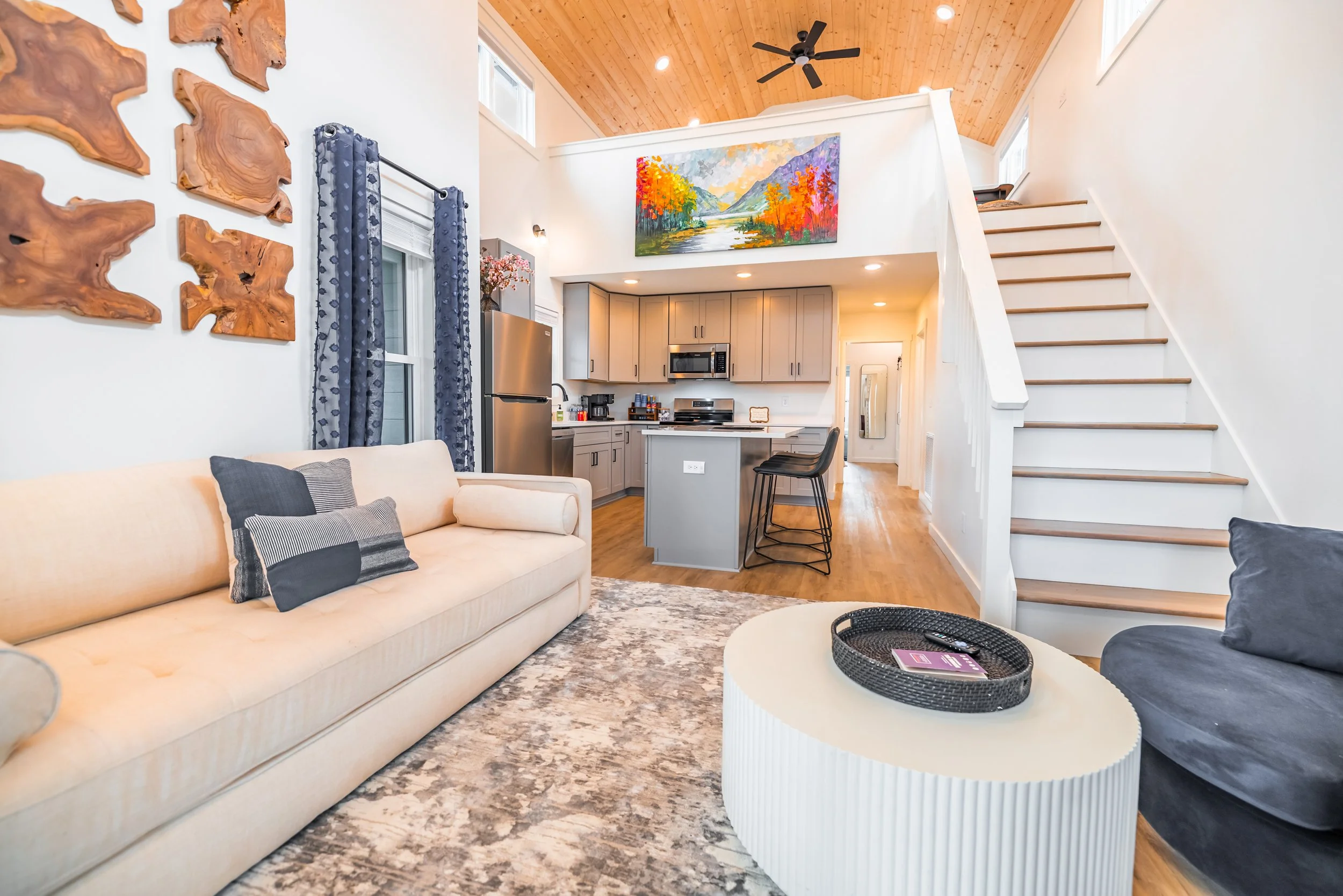 Living room with beige sofa, patterned pillows, dark blue curtains, and a gray accent chair. Open kitchen with gray cabinets, stainless steel refrigerator, and black bar stools at a kitchen island. Stairs leading to a loft with a colorful landscape p