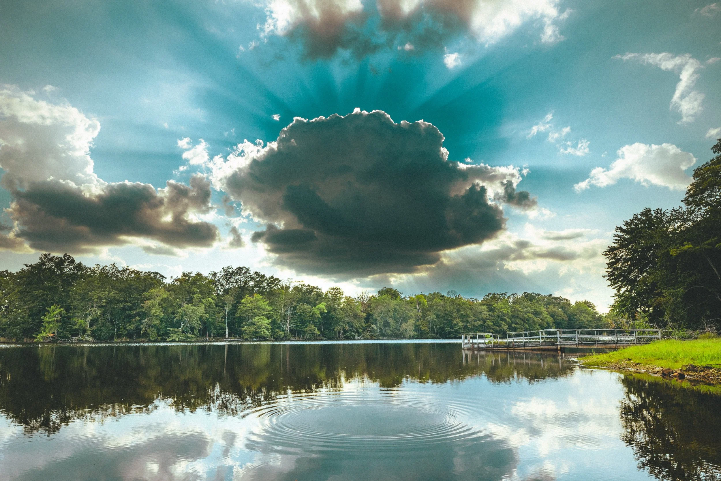 A peaceful lake with calm water reflecting trees and clouds in the sky, with a small dock on the right side and a large cloud centered in the sky