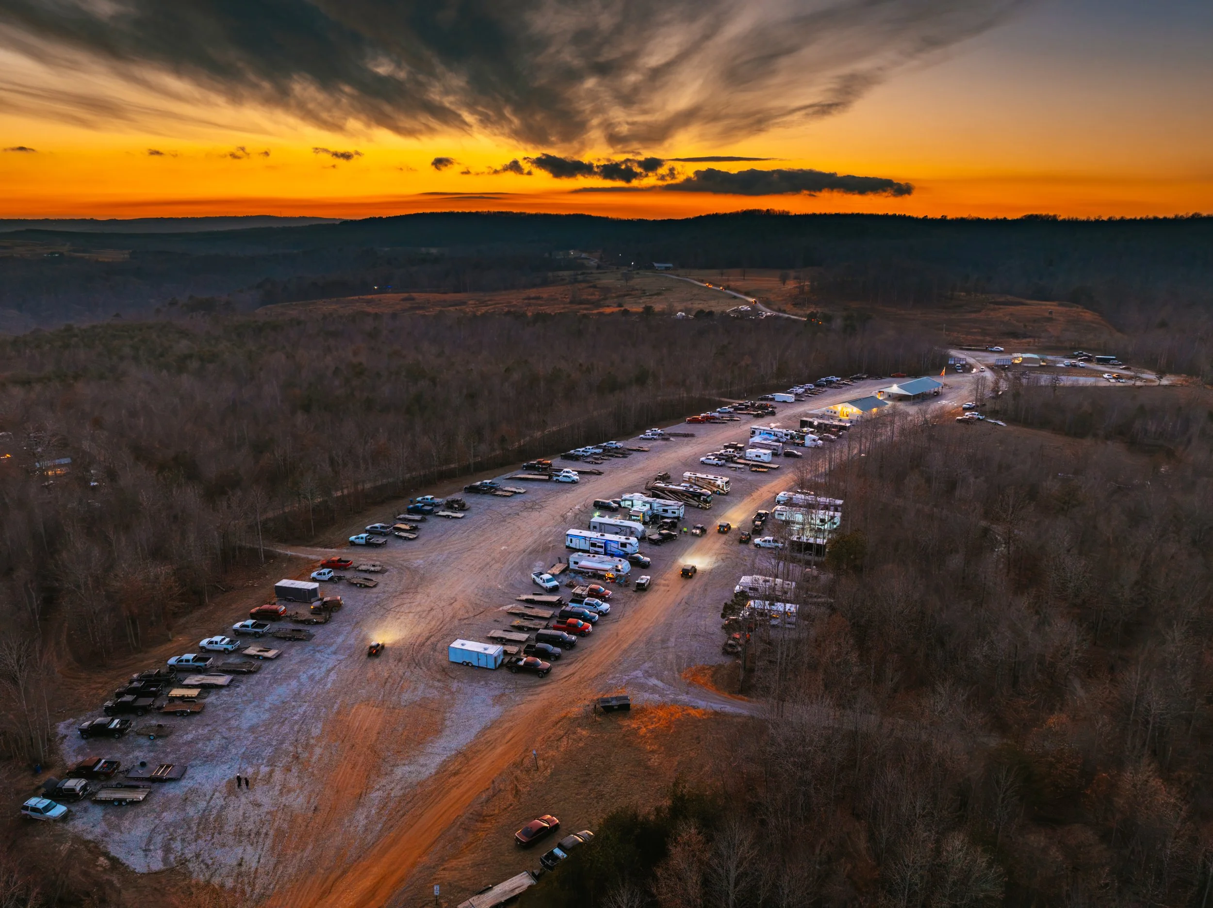 An aerial view of a large outdoor parking area during sunset, with many vehicles including RVs, trucks, and cars parked on a dirt lot surrounded by trees and distant hills.